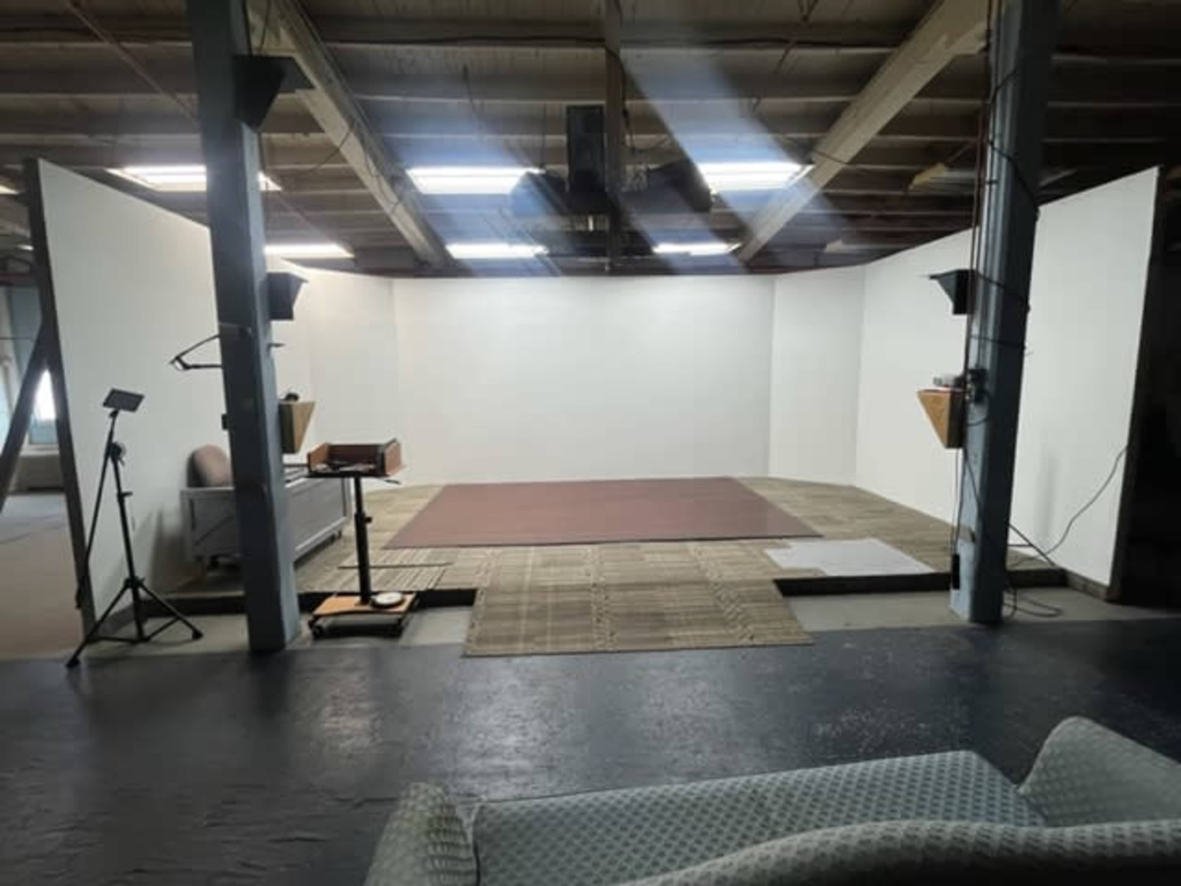 A spacious, empty studio with white walls, a brown rug on the floor, and various filming equipment set up on the sides.