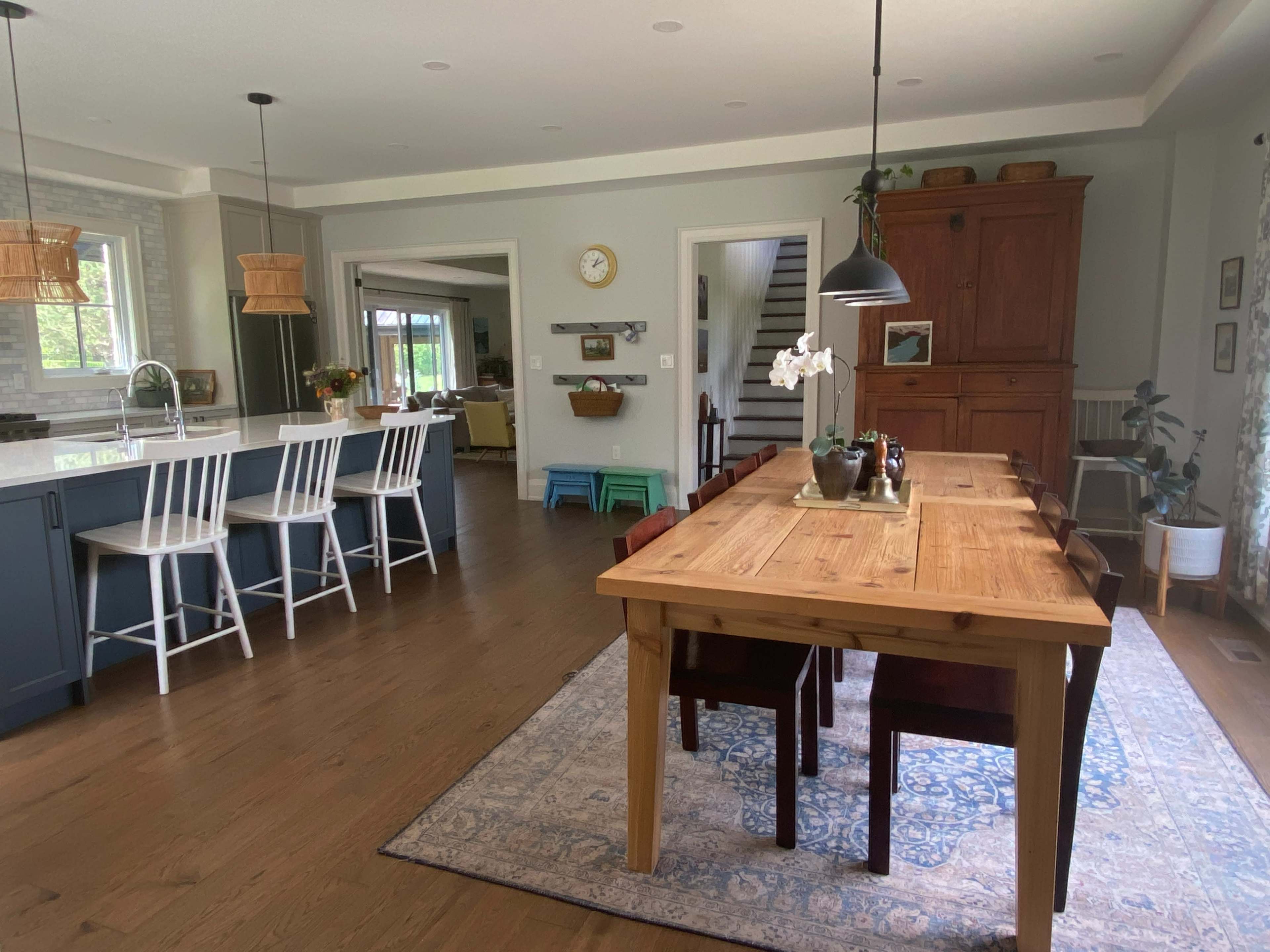 The image shows a spacious kitchen and dining area featuring a wooden dining table, modern bar stools at a kitchen island, and a warm color palette with natural light streaming in.