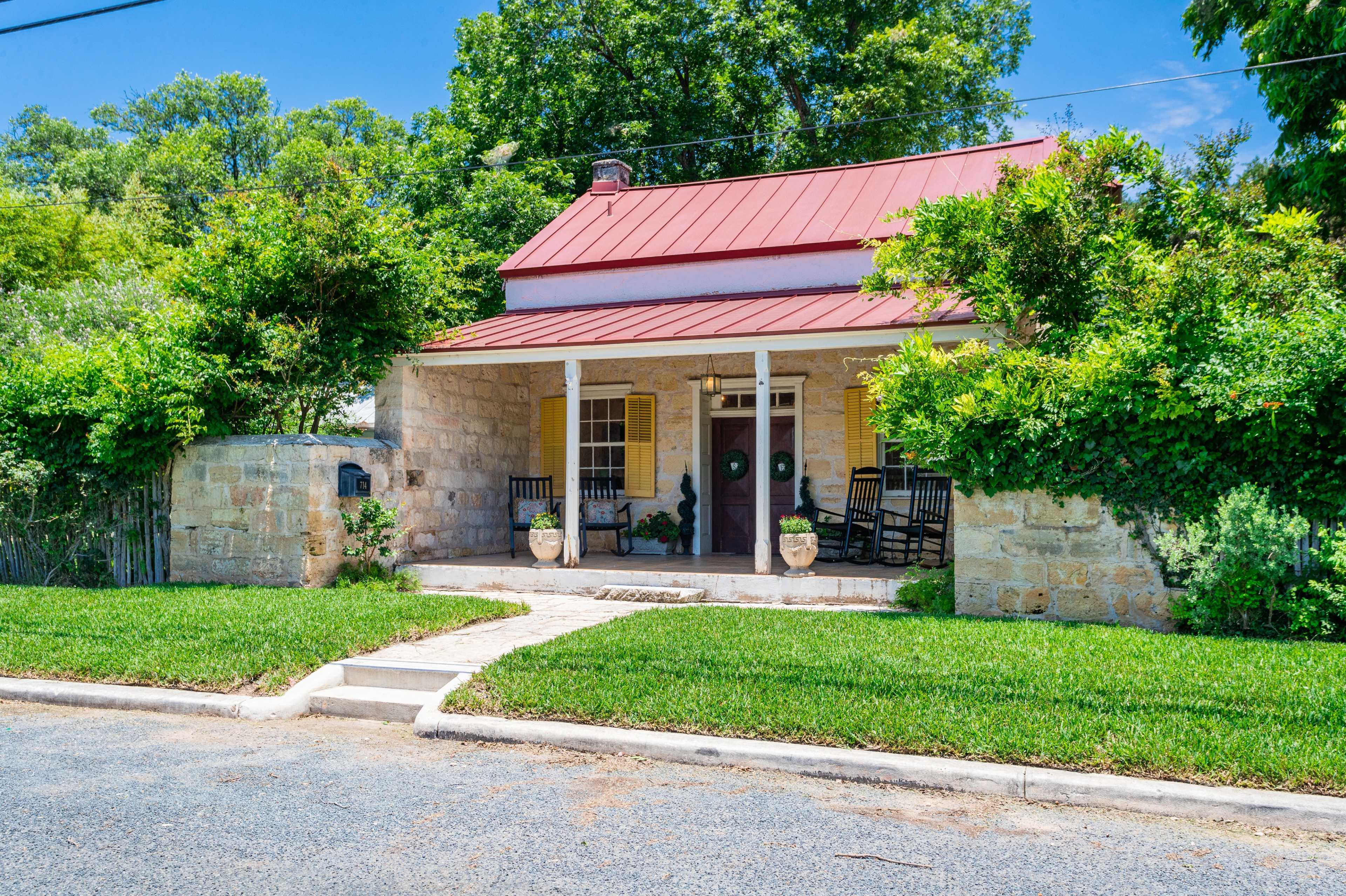 The image shows a stone house with a red metal roof, a front porch with rocking chairs, and potted plants, situated on a grassy yard beside a street.