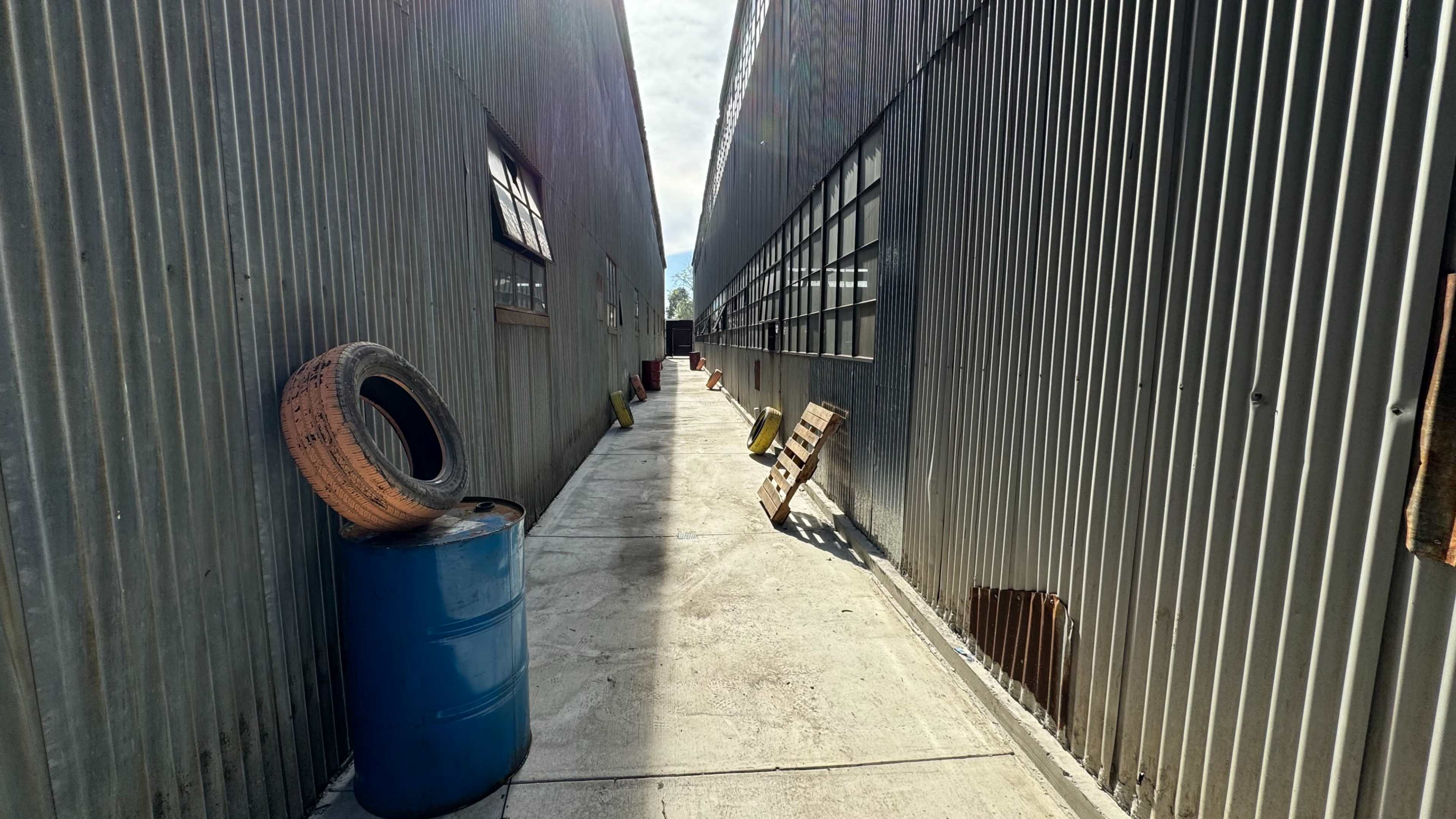 A narrow passage between two corrugated metal buildings, lined with a blue barrel and a tire, with wooden pallets placed along the wall.