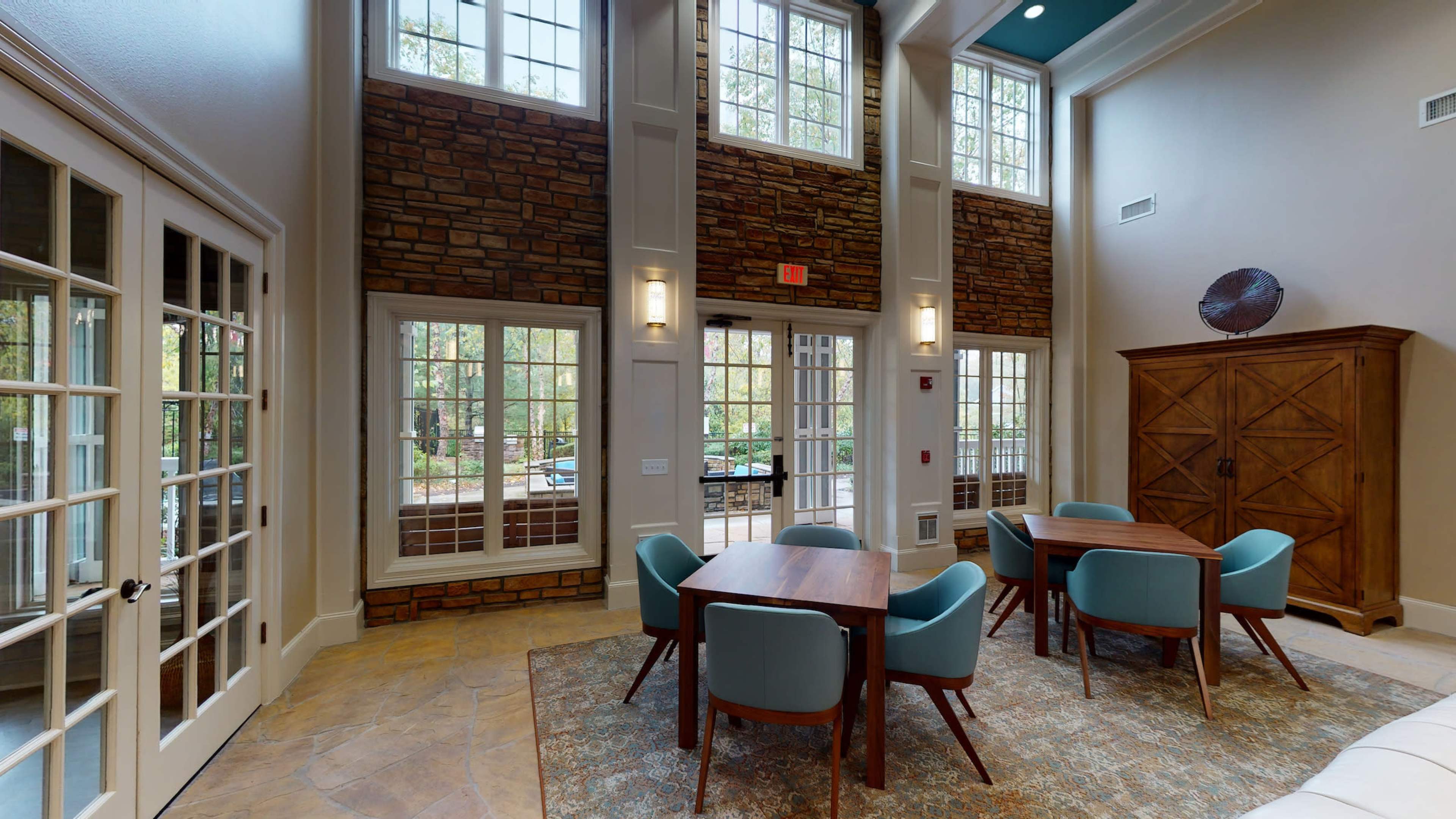 A bright room with large windows, featuring several wooden tables and blue chairs on a patterned rug.
