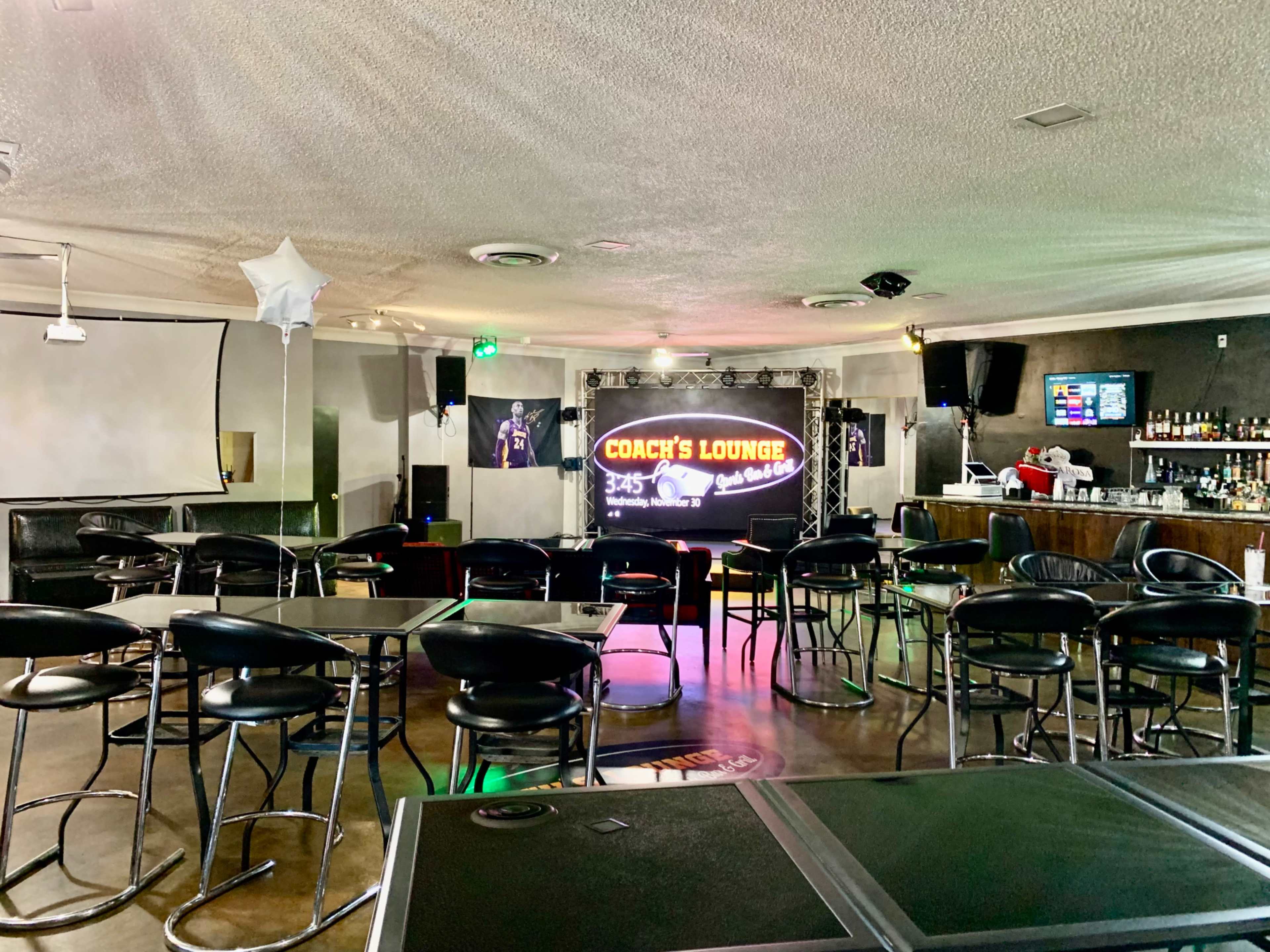 A lounge area with black bar stools and tables, featuring a performance stage with a lighted sign that reads "Coach's Lounge."