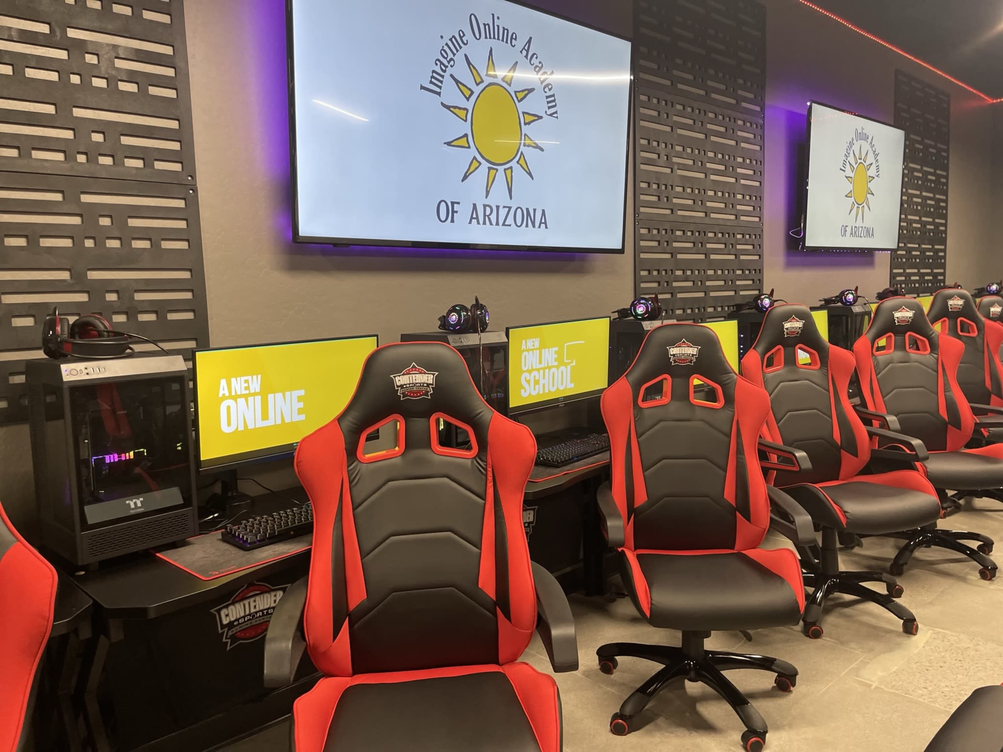 A modern classroom setting with several gaming chairs and computer stations, featuring a prominent logo for "Imagine Online Academy of Arizona" on a screen at the front.