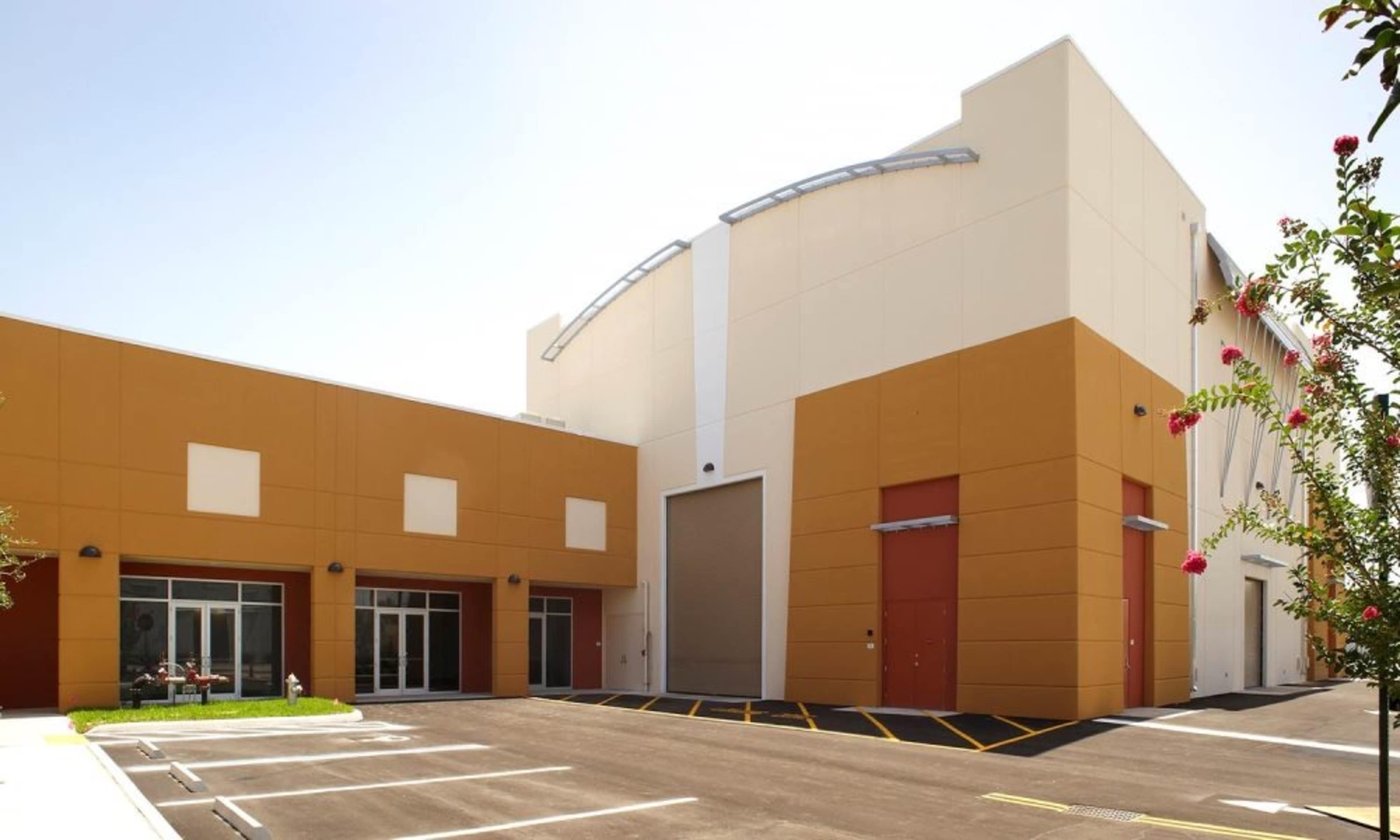 The image shows a modern industrial building with a large parking area, featuring a combination of orange and beige exterior walls.
