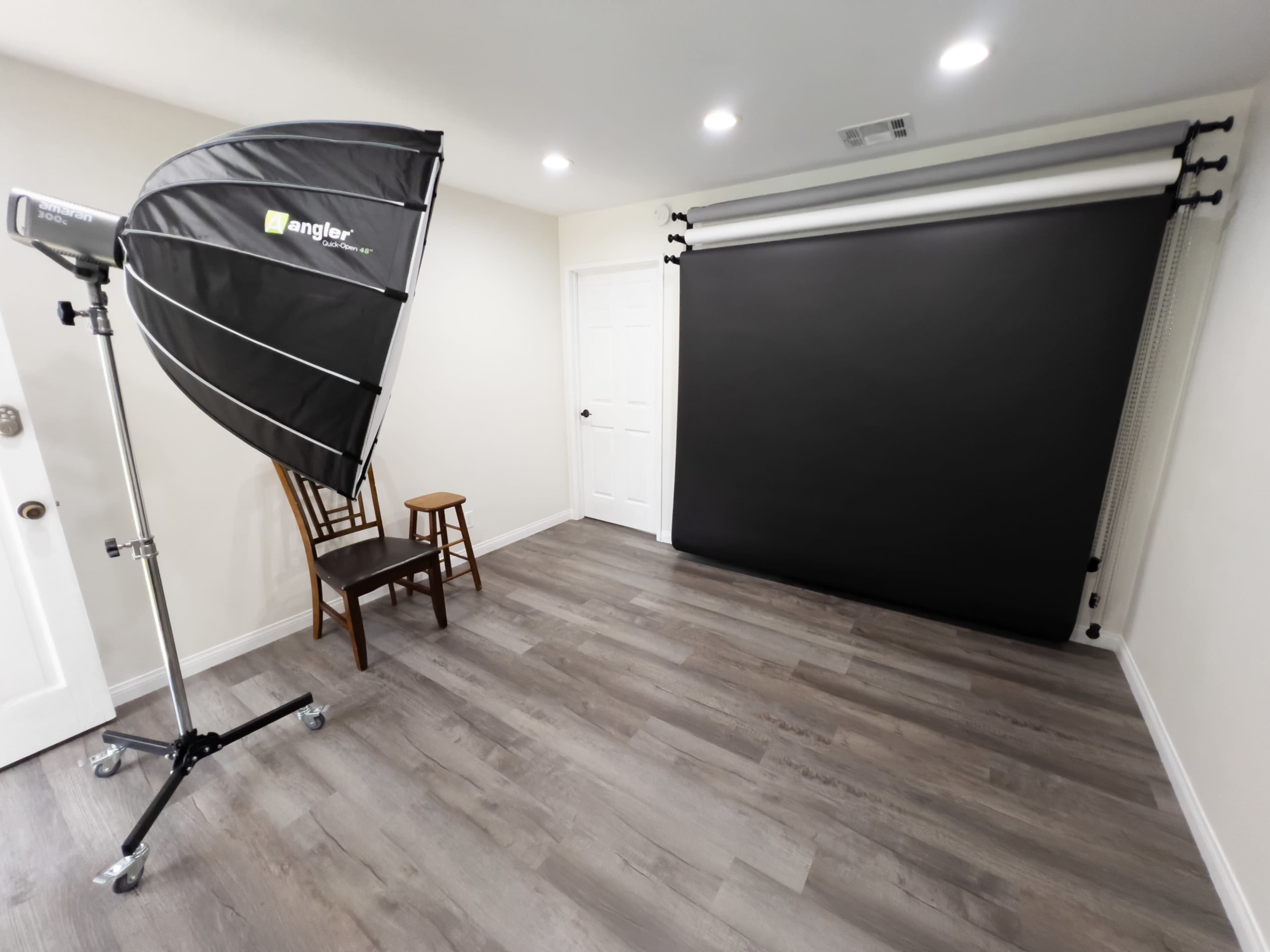 A photography studio with a black backdrop, a light source on a stand, a chair, and wooden flooring.