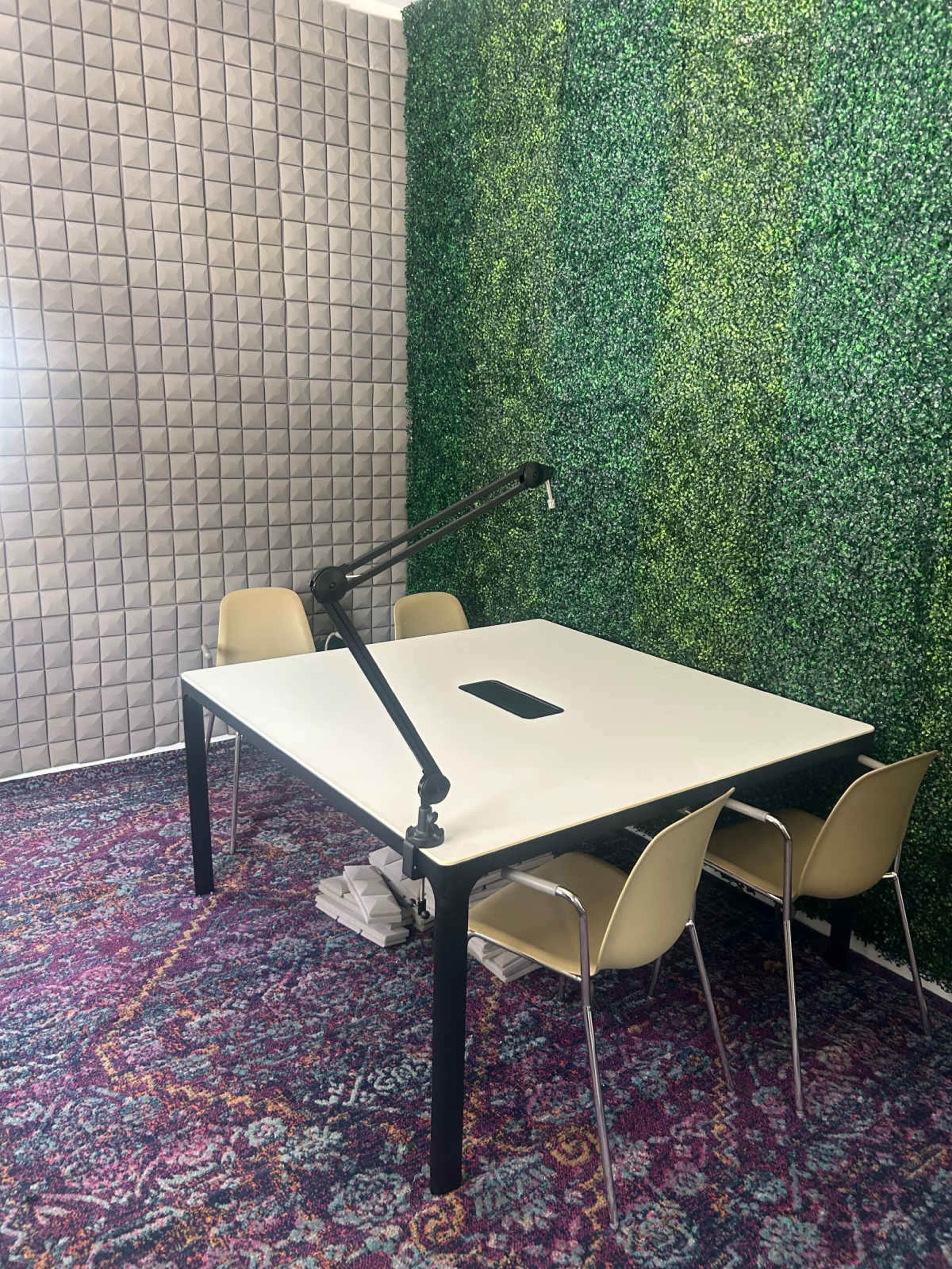 The image shows a small meeting room featuring a white table with a microphone arm, four beige chairs, and walls covered with green artificial foliage and acoustic panels.