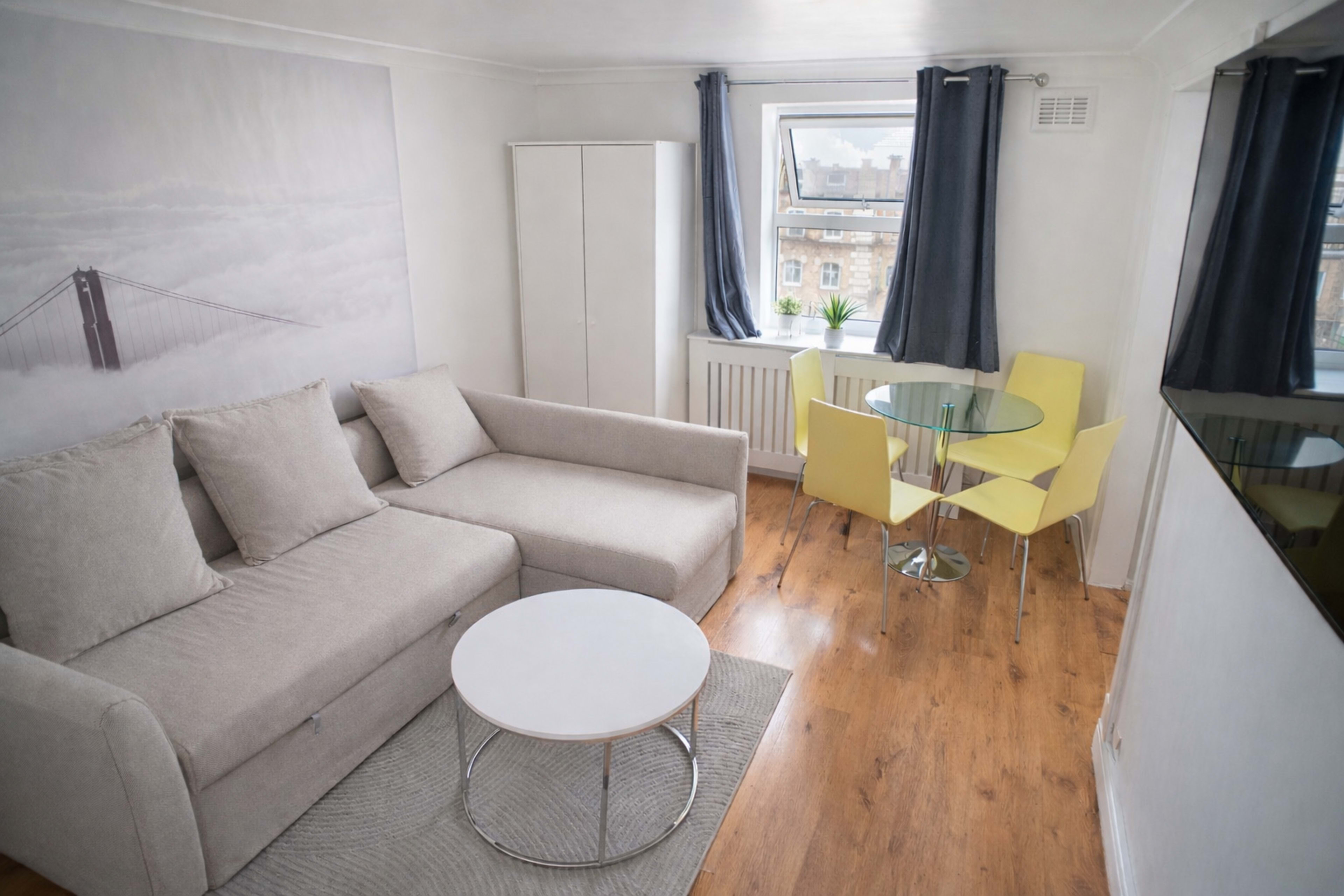 The image shows a small living area with a grey sofa, a round white coffee table, and a glass dining table with four yellow chairs, complemented by a window and a wall featuring a picture of a bridge.