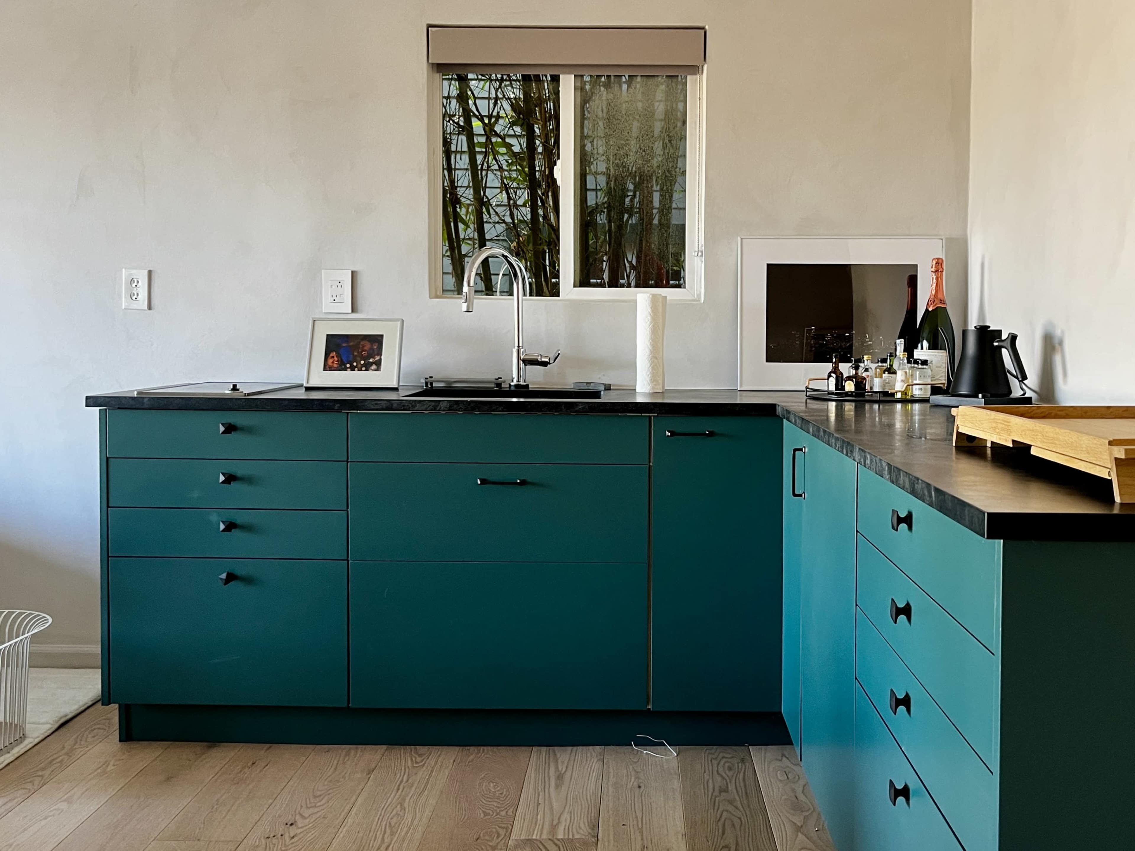 The image shows a modern kitchen with teal cabinets, a sink, a countertop, and a window facing an outdoor view.