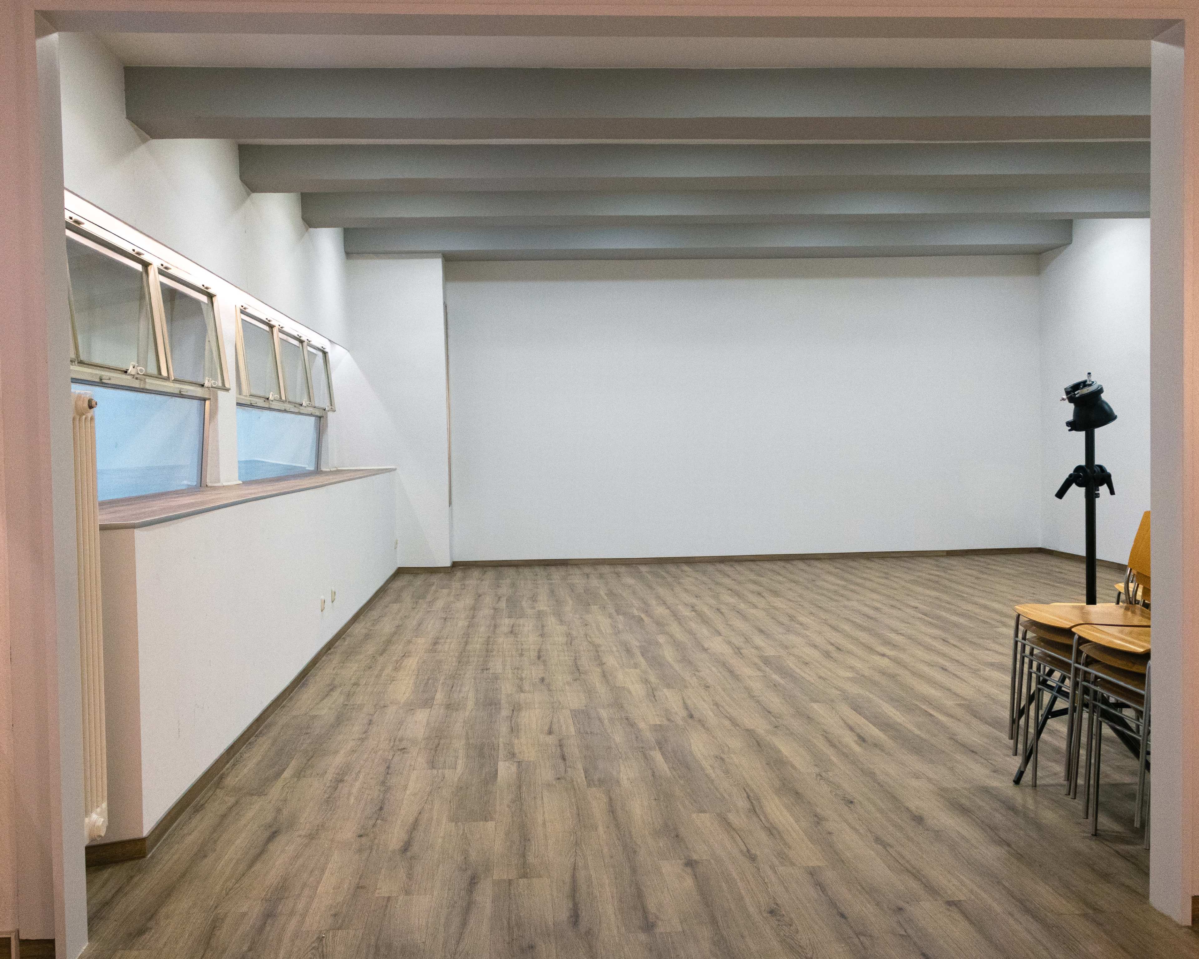 The image shows a spacious, empty room with wooden flooring, large windows, and stacked chairs against one wall.