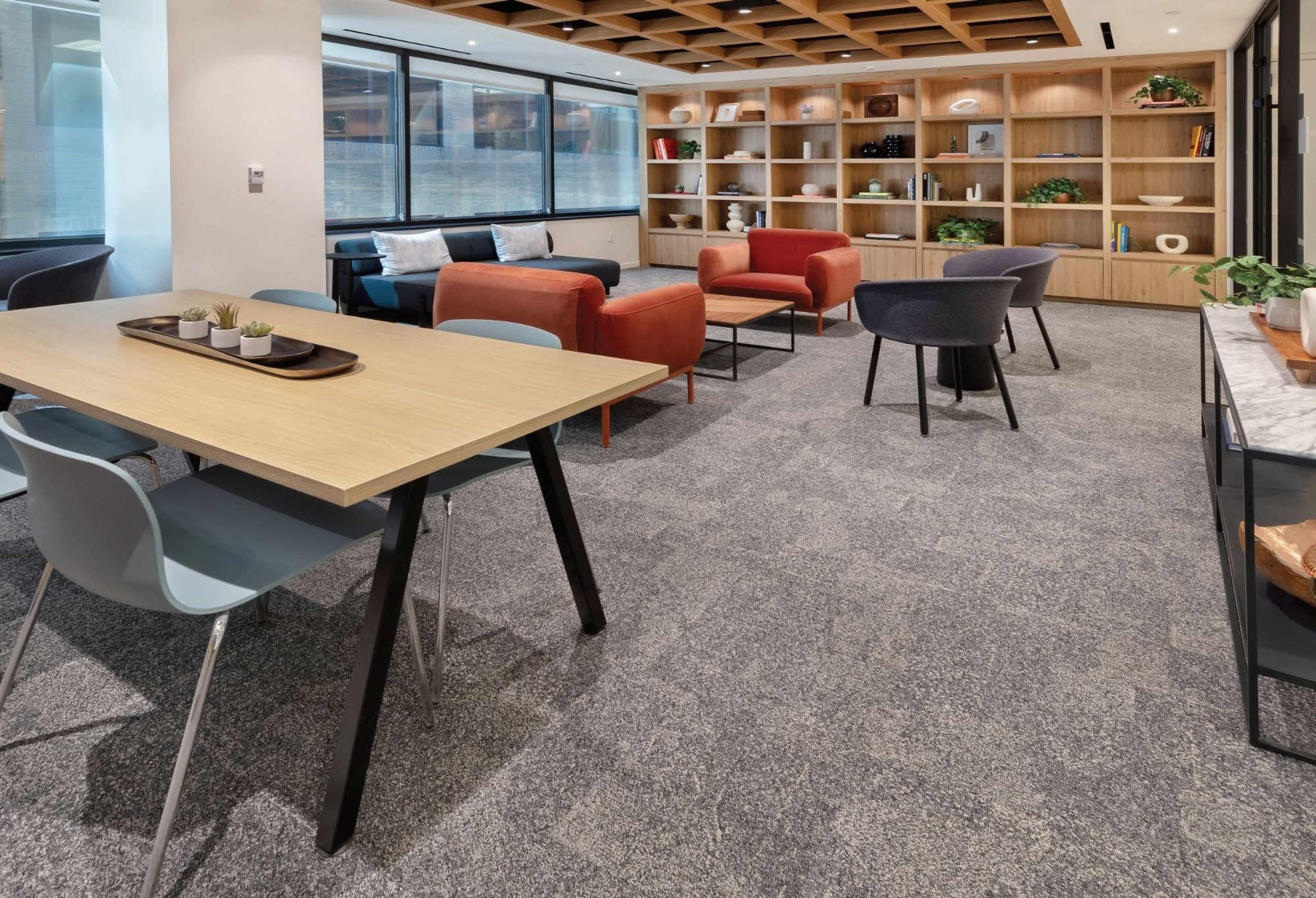 An open office lounge features a wooden table with chairs, a bookshelf filled with decor, and various seating arrangements including red and gray armchairs.