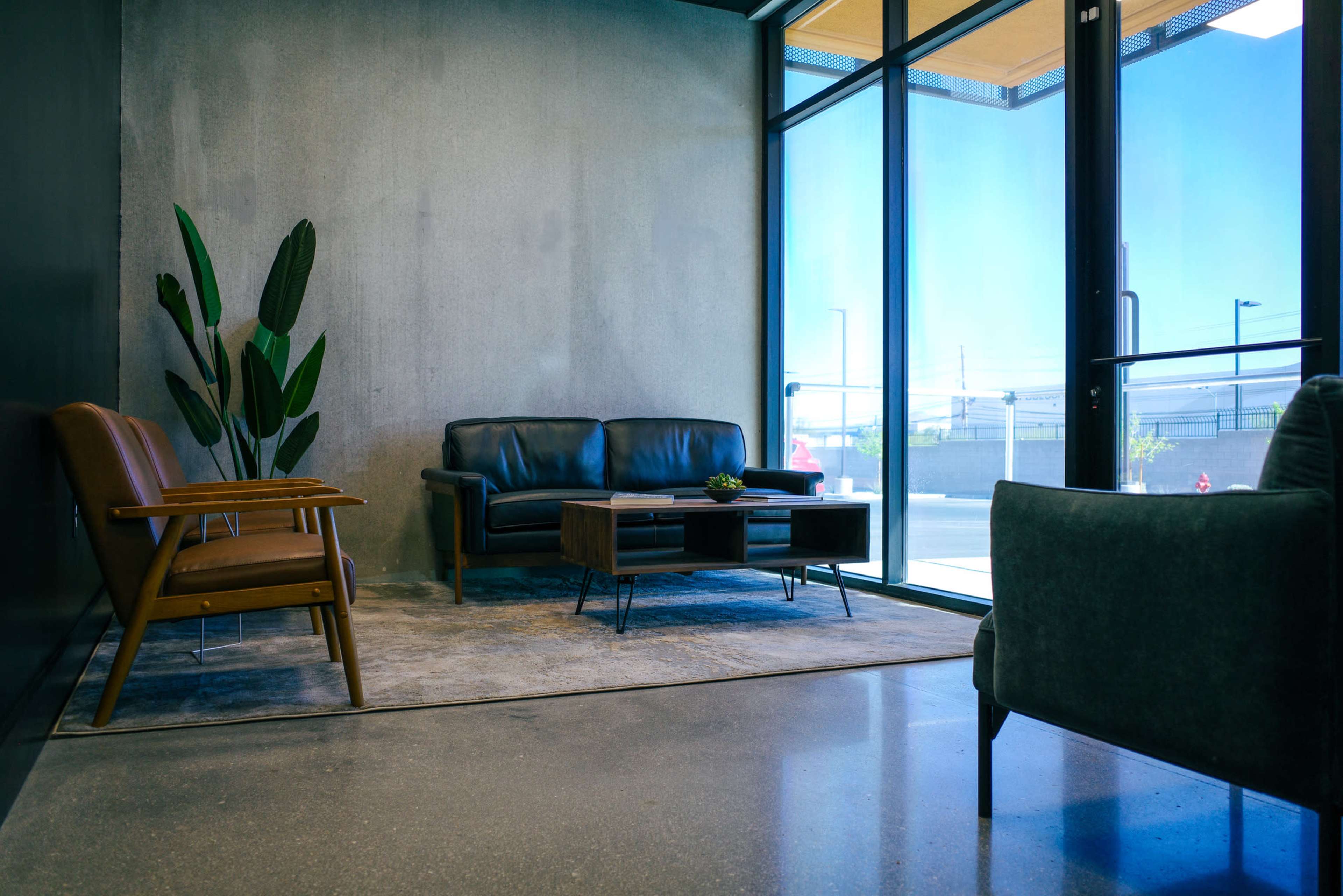 The image shows a minimalist lounge area with a black leather couch, a wooden chair, and large windows allowing natural light to enter.