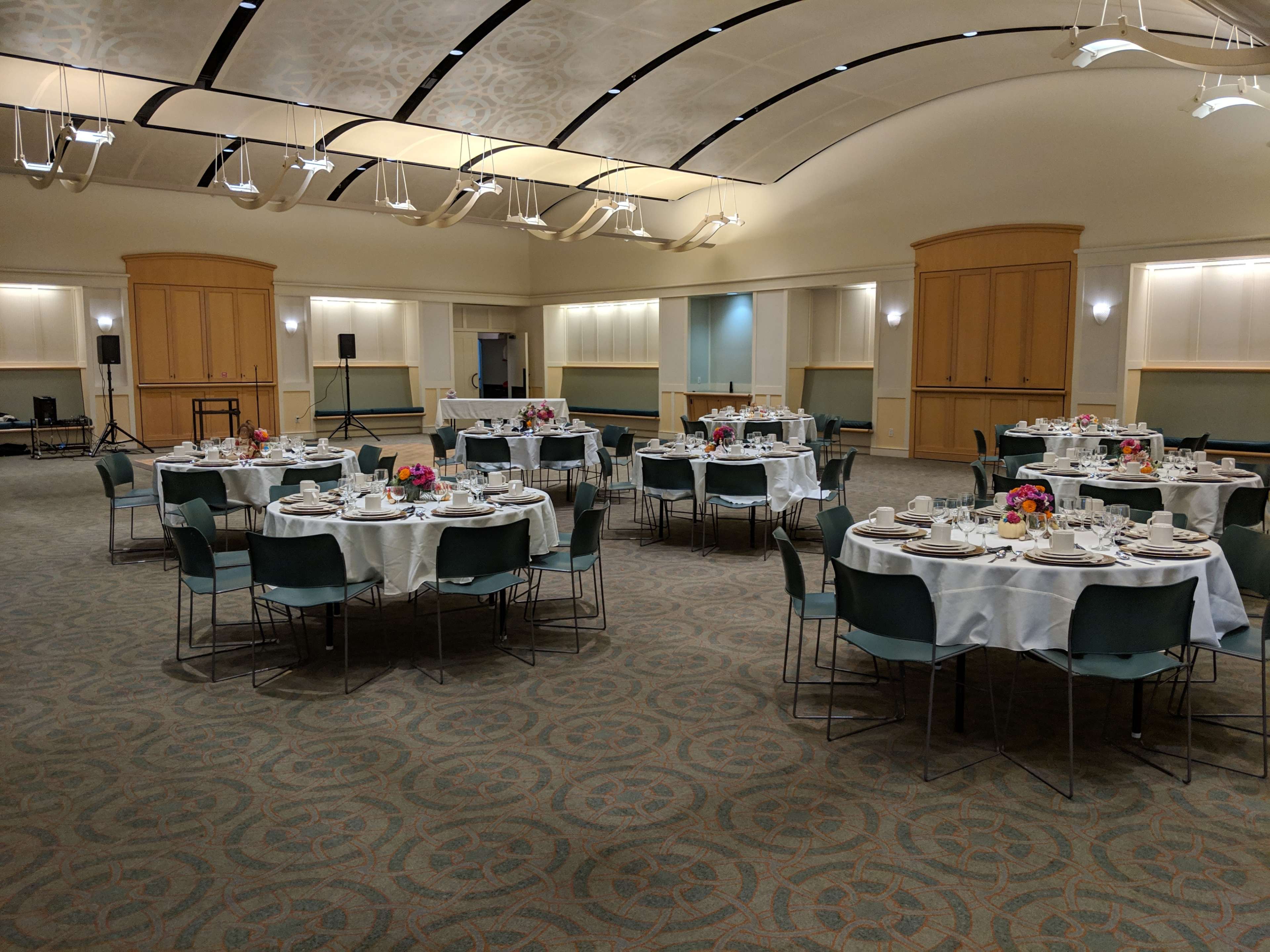 A banquet hall is set with several round tables adorned with white tablecloths and centerpieces of flowers, arranged for a formal dining event.