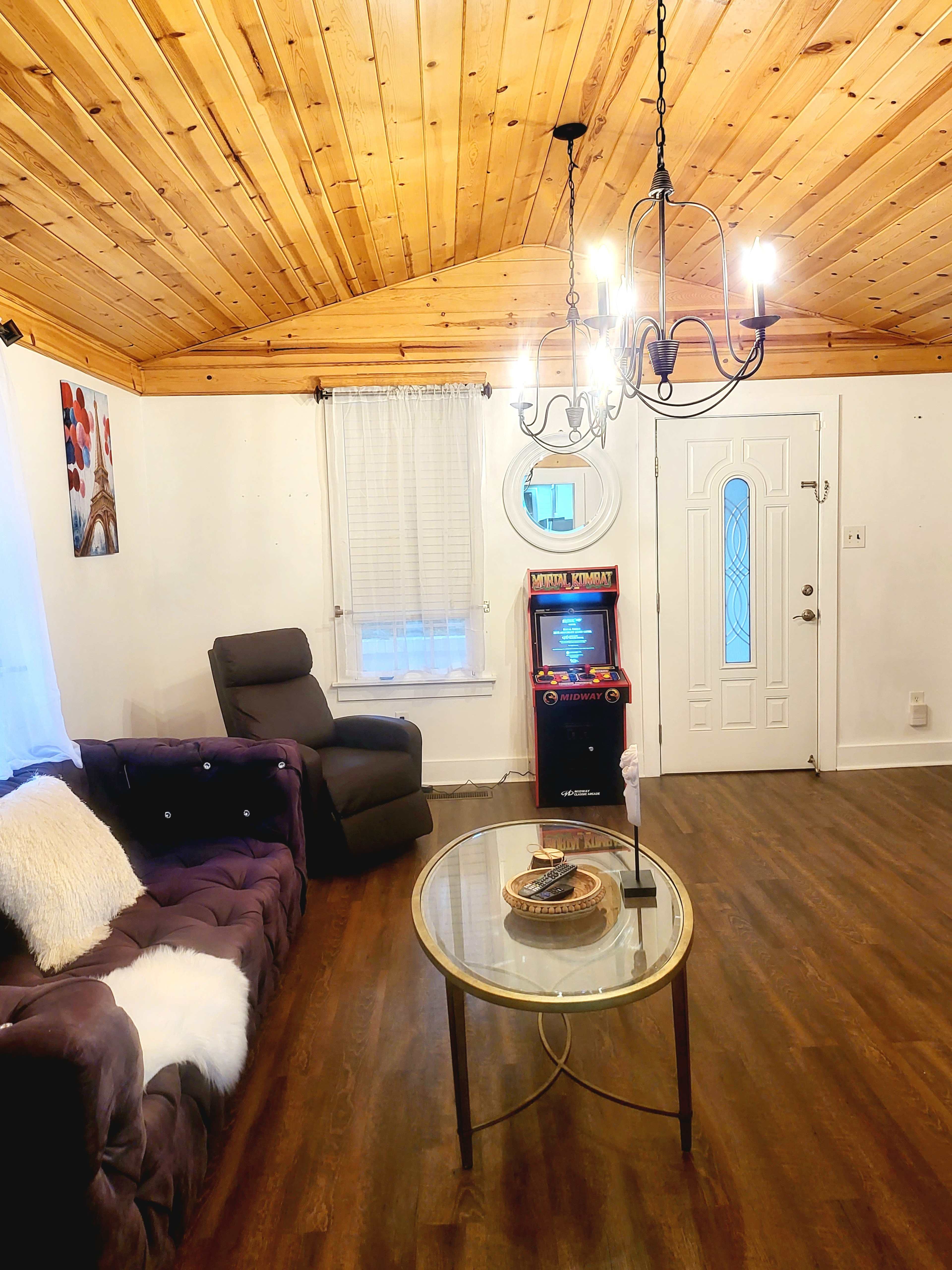 The living room features a wooden ceiling, a plush sofa, a glass coffee table, and an arcade machine by the door.