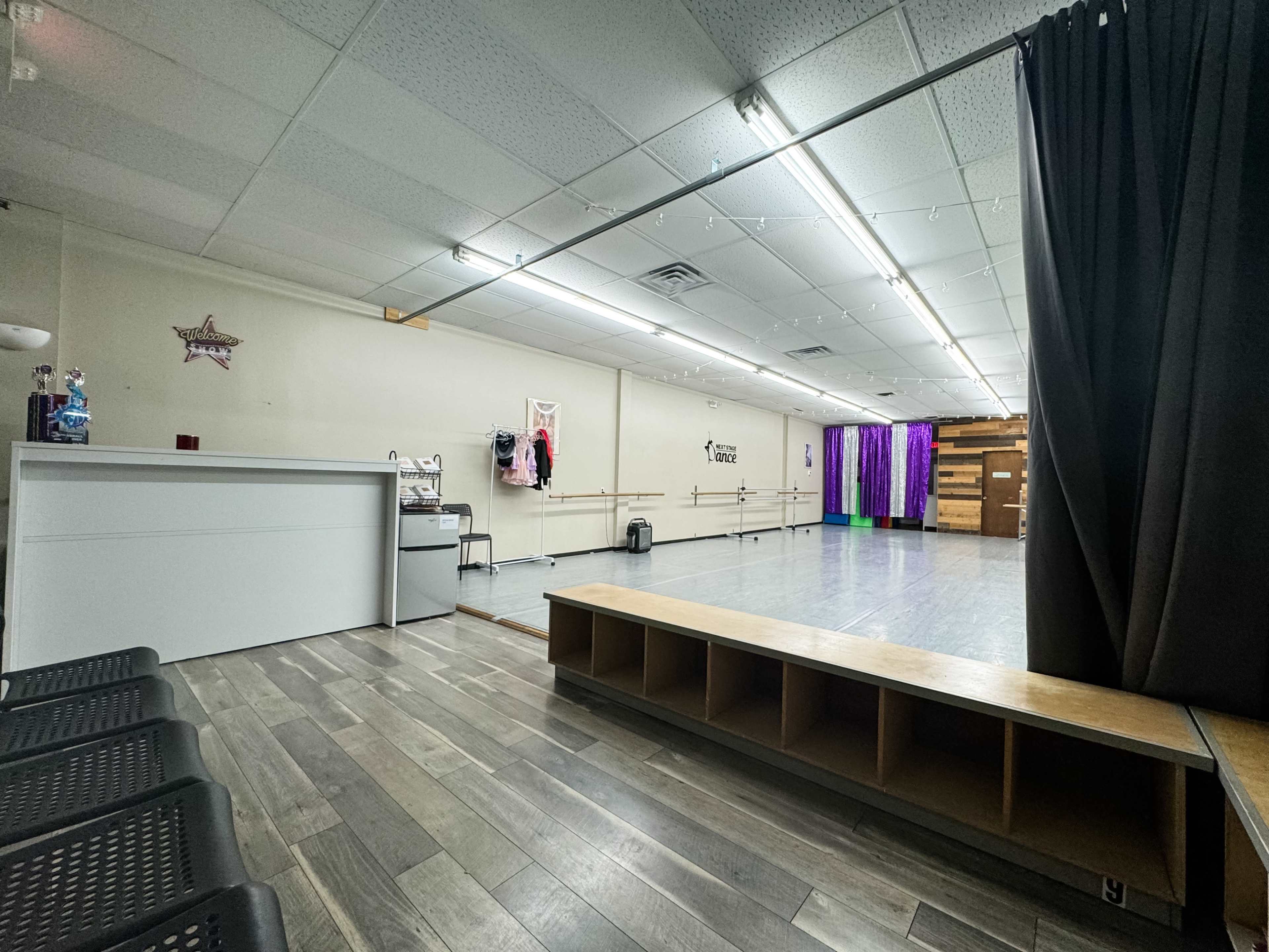 The image shows a spacious dance studio with wooden flooring, mirrors, and a seating area along one side.