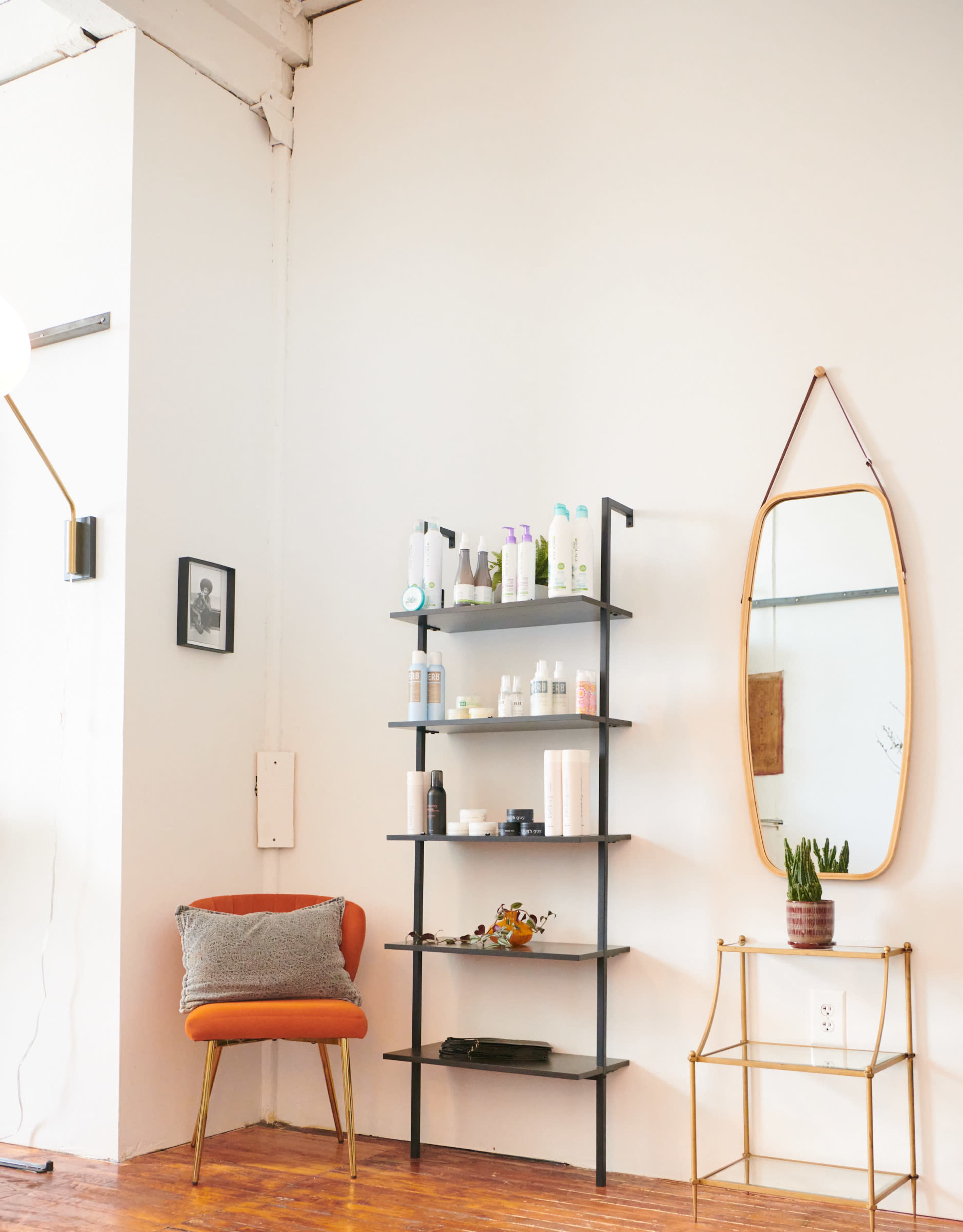 A brown chair with a cushion is positioned beside a black metal shelf filled with various skincare products, against a white wall adorned with a mirror and a small plant.