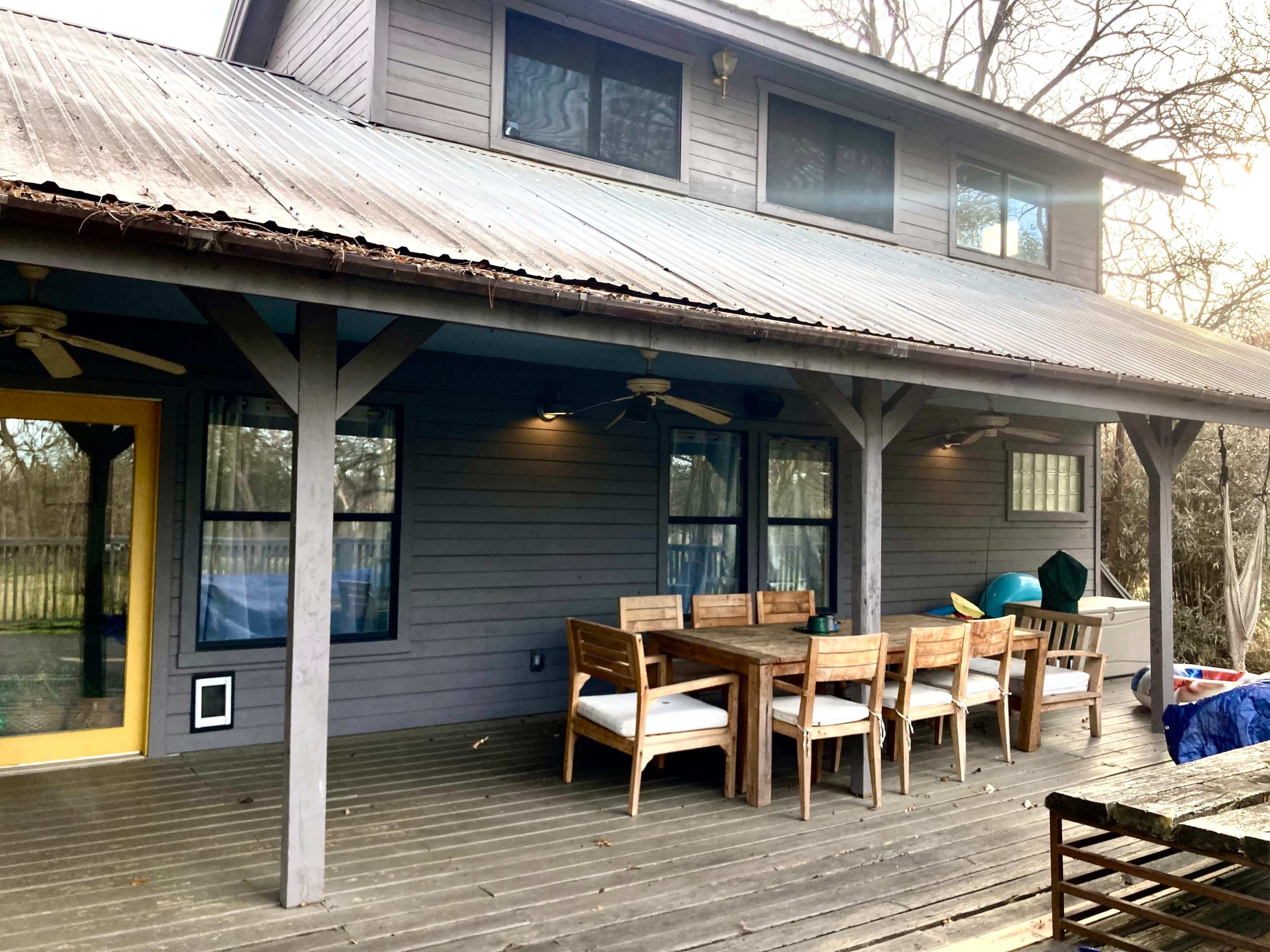 A wooden deck features a large dining table and chairs under a covered area of a two-story house.