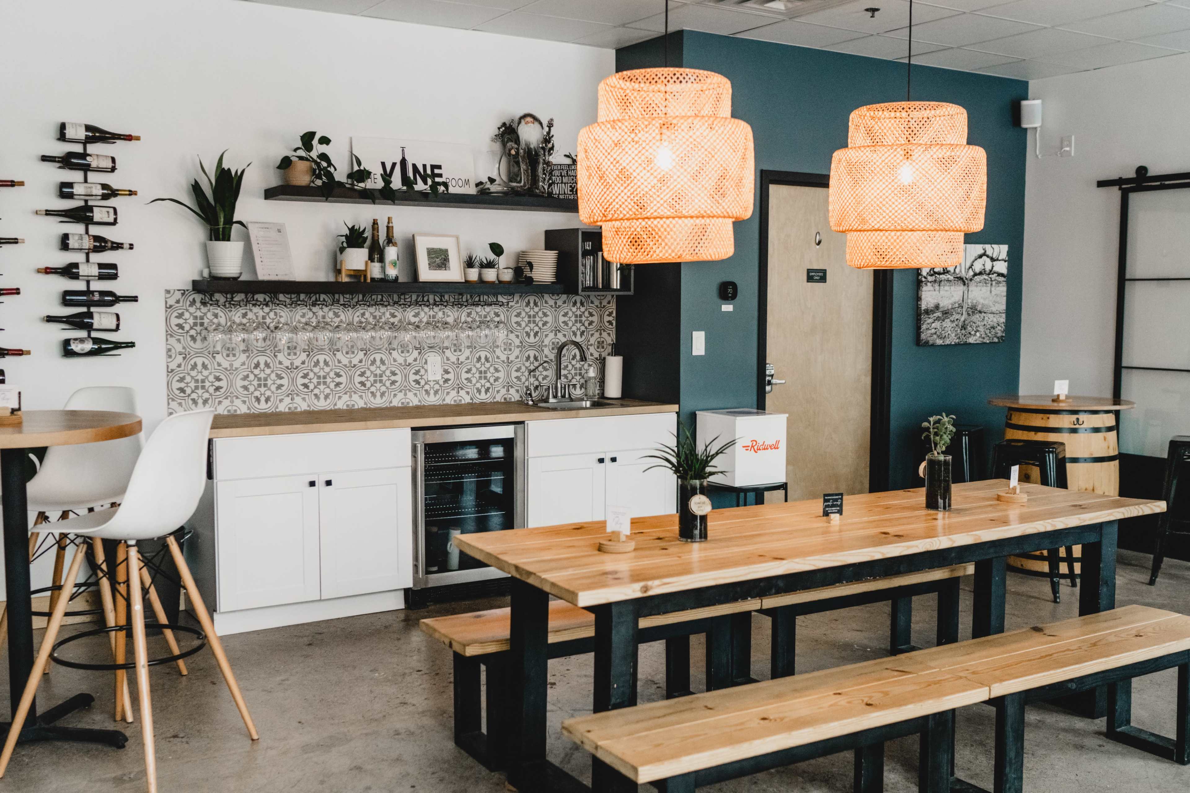 A modern interior of a spacious room with wooden tables, a white kitchen area, and pendant lighting hanging above.