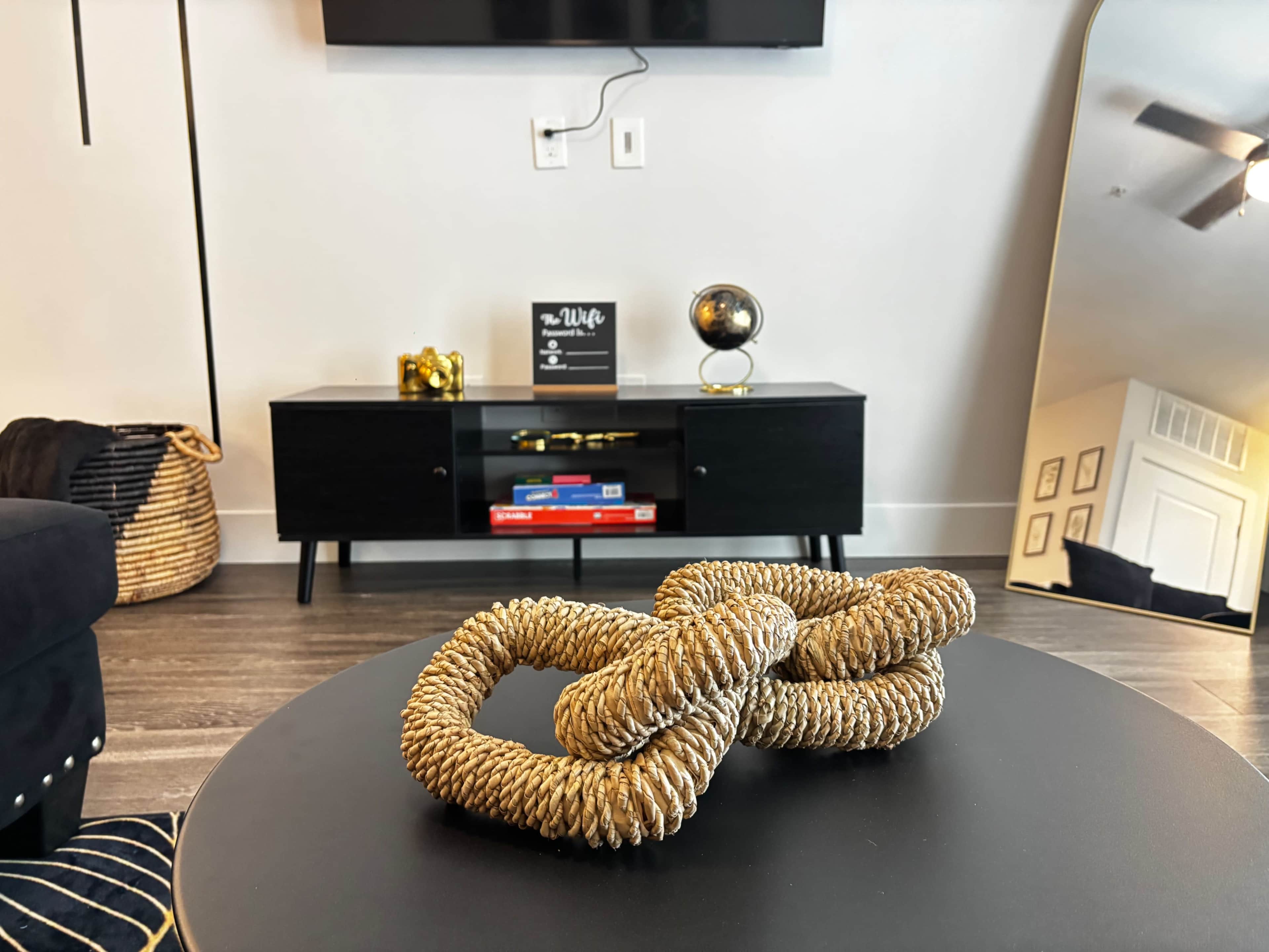 The image shows a modern living room with a black coffee table featuring a decorative woven knot, a black entertainment unit, and a large mirror reflecting part of the space.