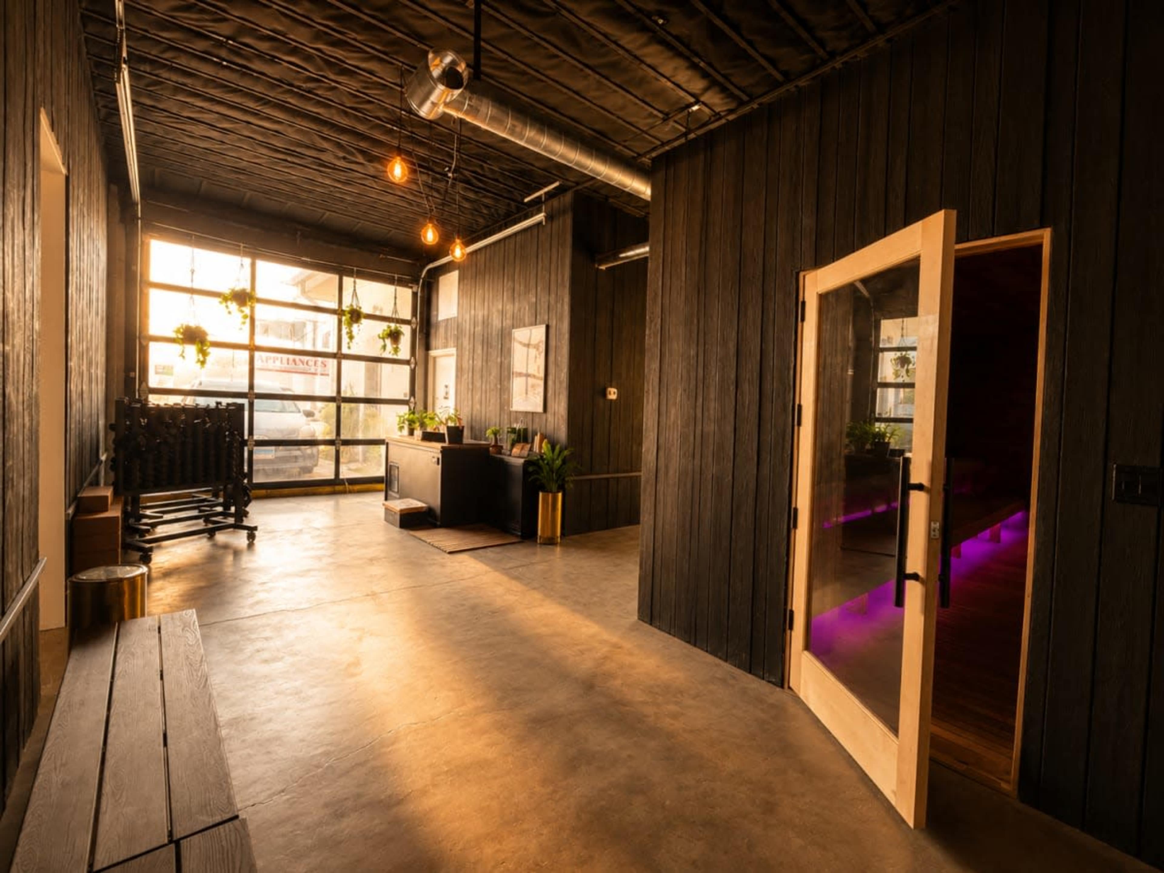 The image shows a modern interior space with wooden walls, concrete flooring, and large windows, featuring a reception area and a doorway leading to another room.