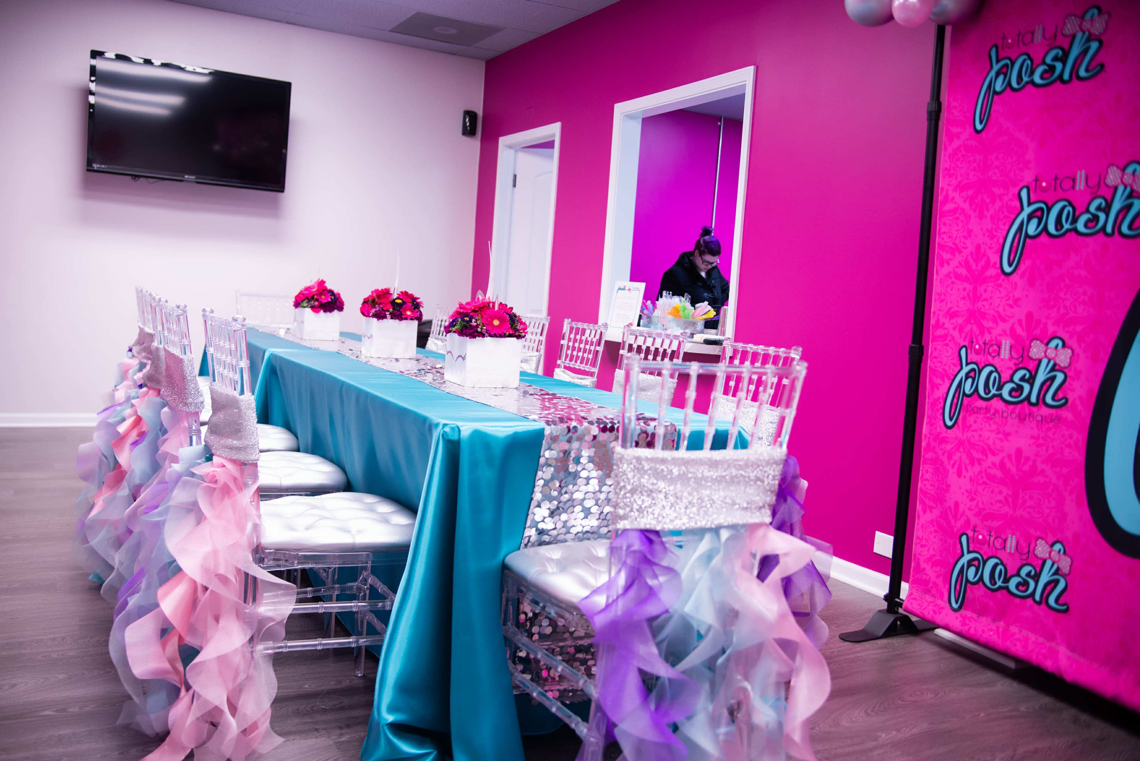 A colorful event space features a long table set with a turquoise tablecloth, white chairs adorned with ruffled covers, and floral centerpieces.