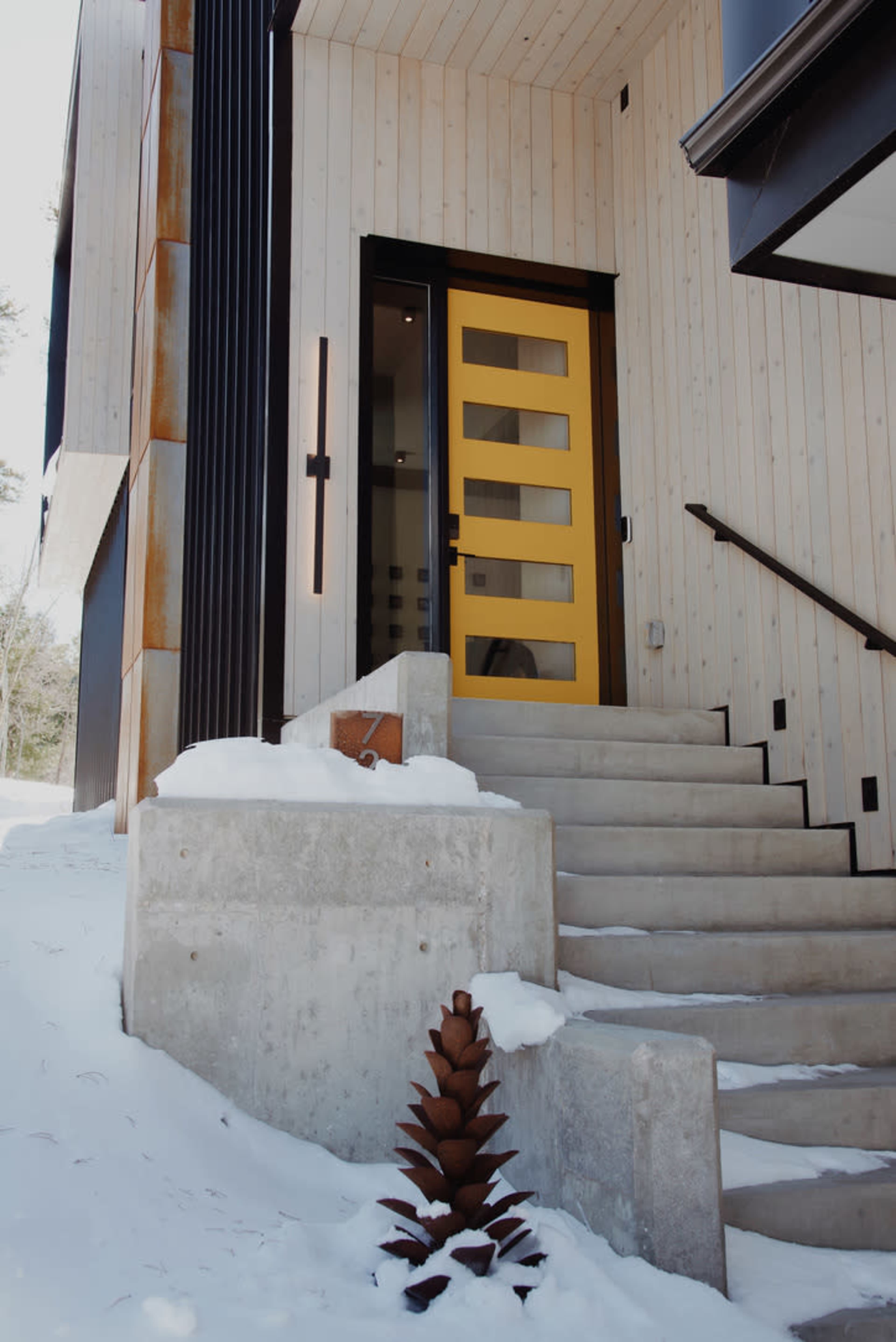The image shows a modern building entrance with a bright yellow door, concrete steps, and a snow-covered landscape.