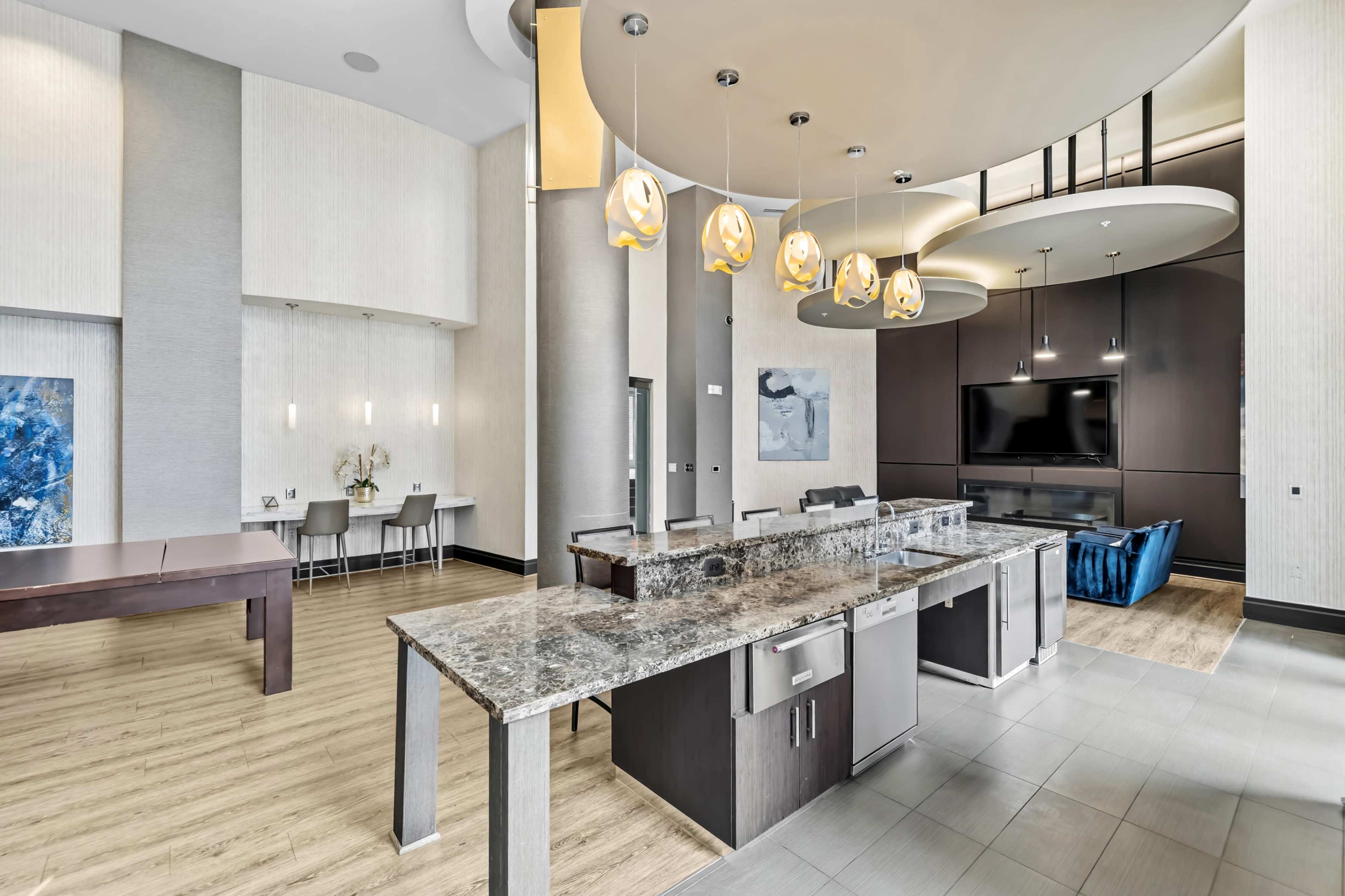 The image shows a contemporary dining and living area featuring a large kitchen island, modern light fixtures, and a wall-mounted television.