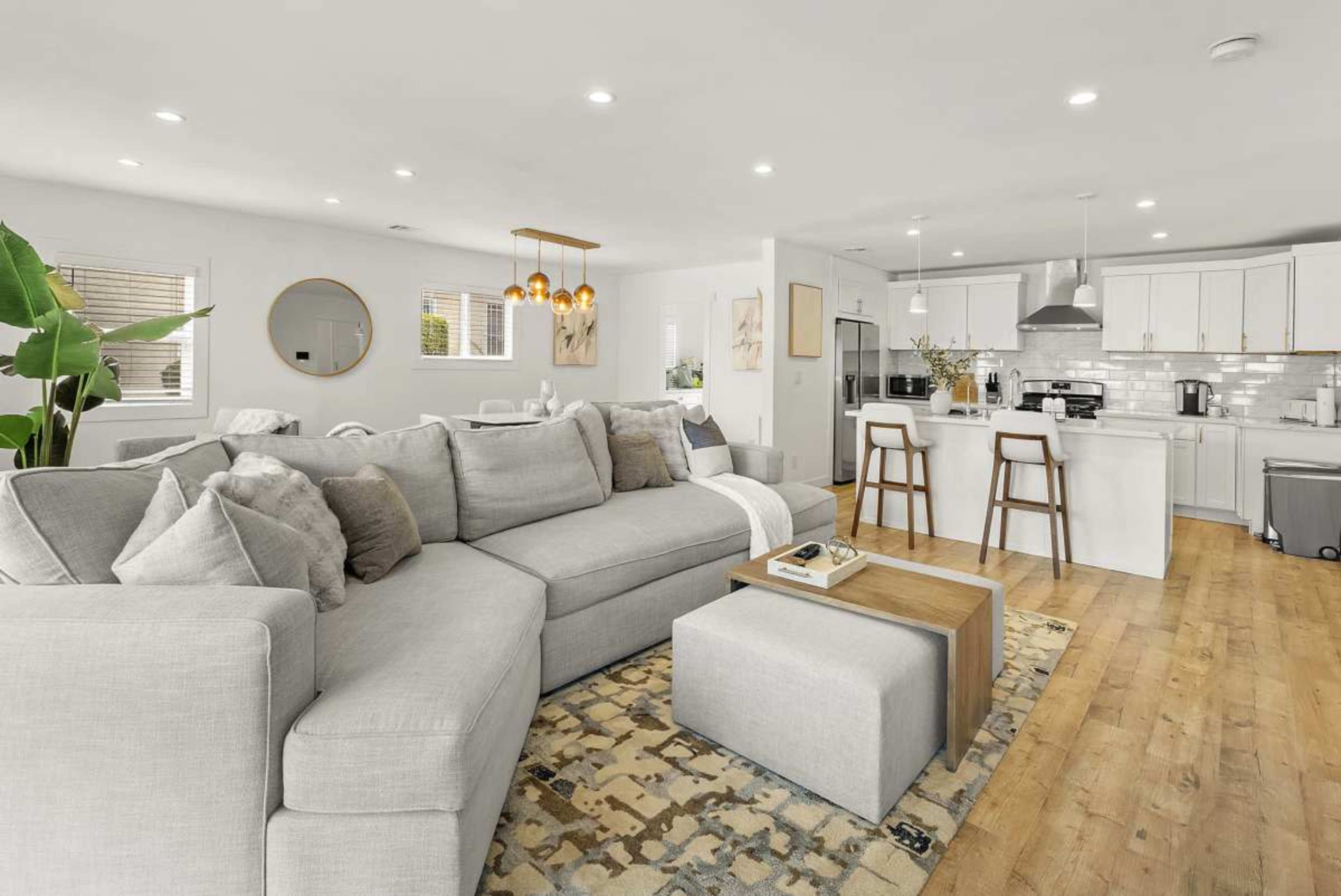 The image depicts a modern, open-concept living space with a large gray sectional sofa, a wooden coffee table, and a kitchen area featuring white cabinetry and bar stools.