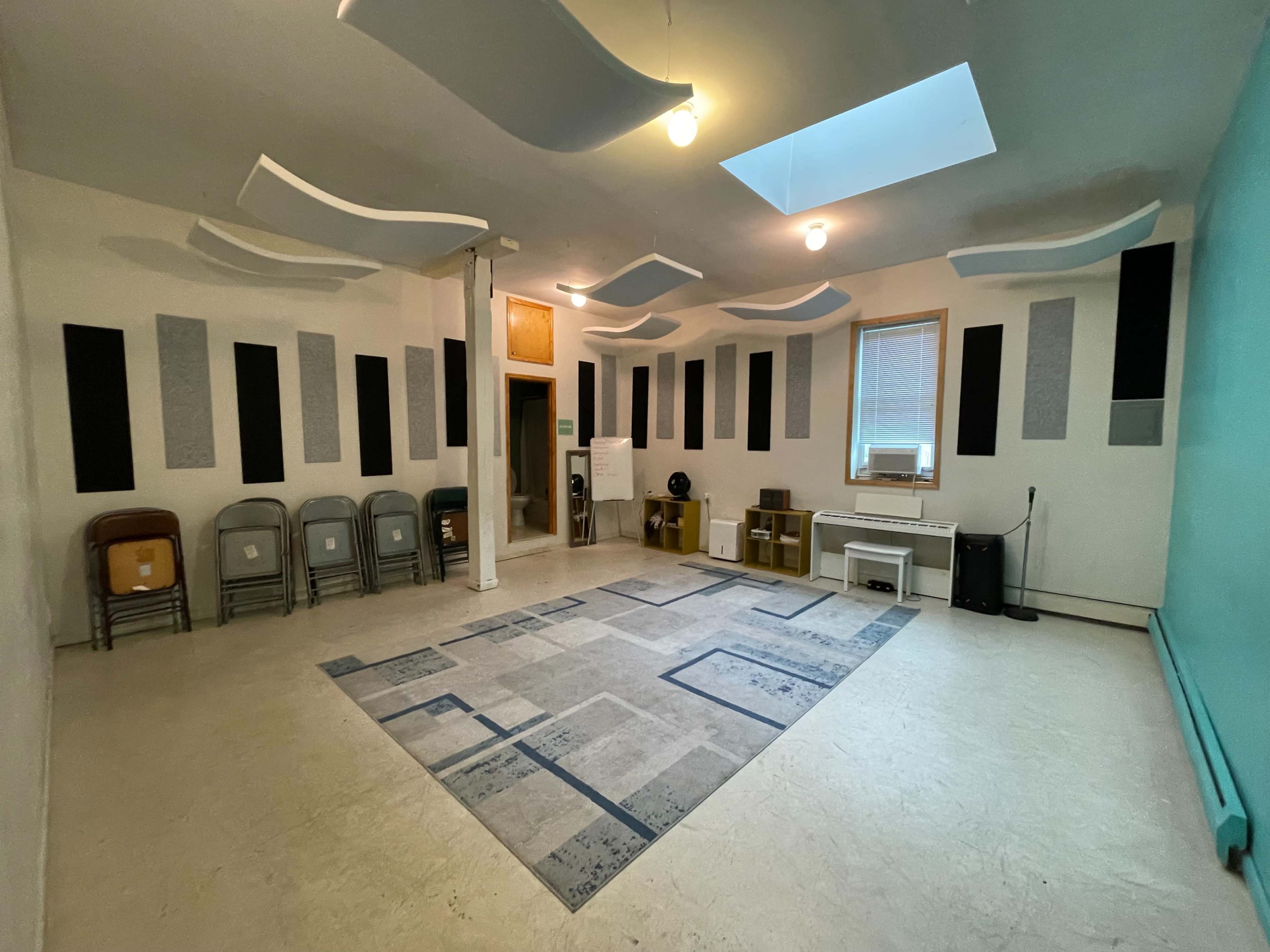The image shows a spacious room with a light-colored floor, various sound-absorbing panels on the walls, a patterned rug in the center, and a set of folding chairs arranged on one side.
