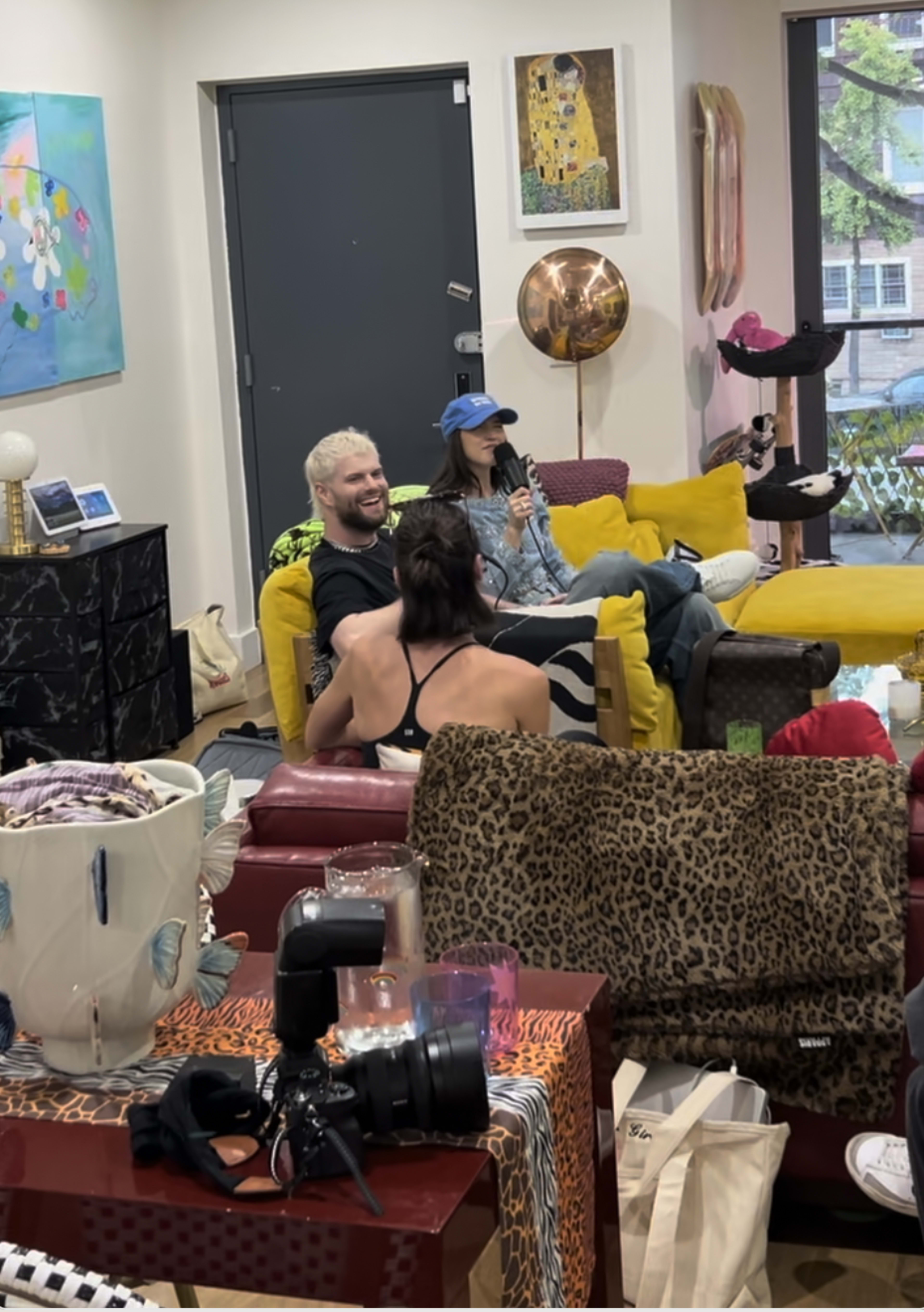 Two people sit in a brightly decorated living room, one on a yellow couch and another on a red couch, while a third person speaks in the background.