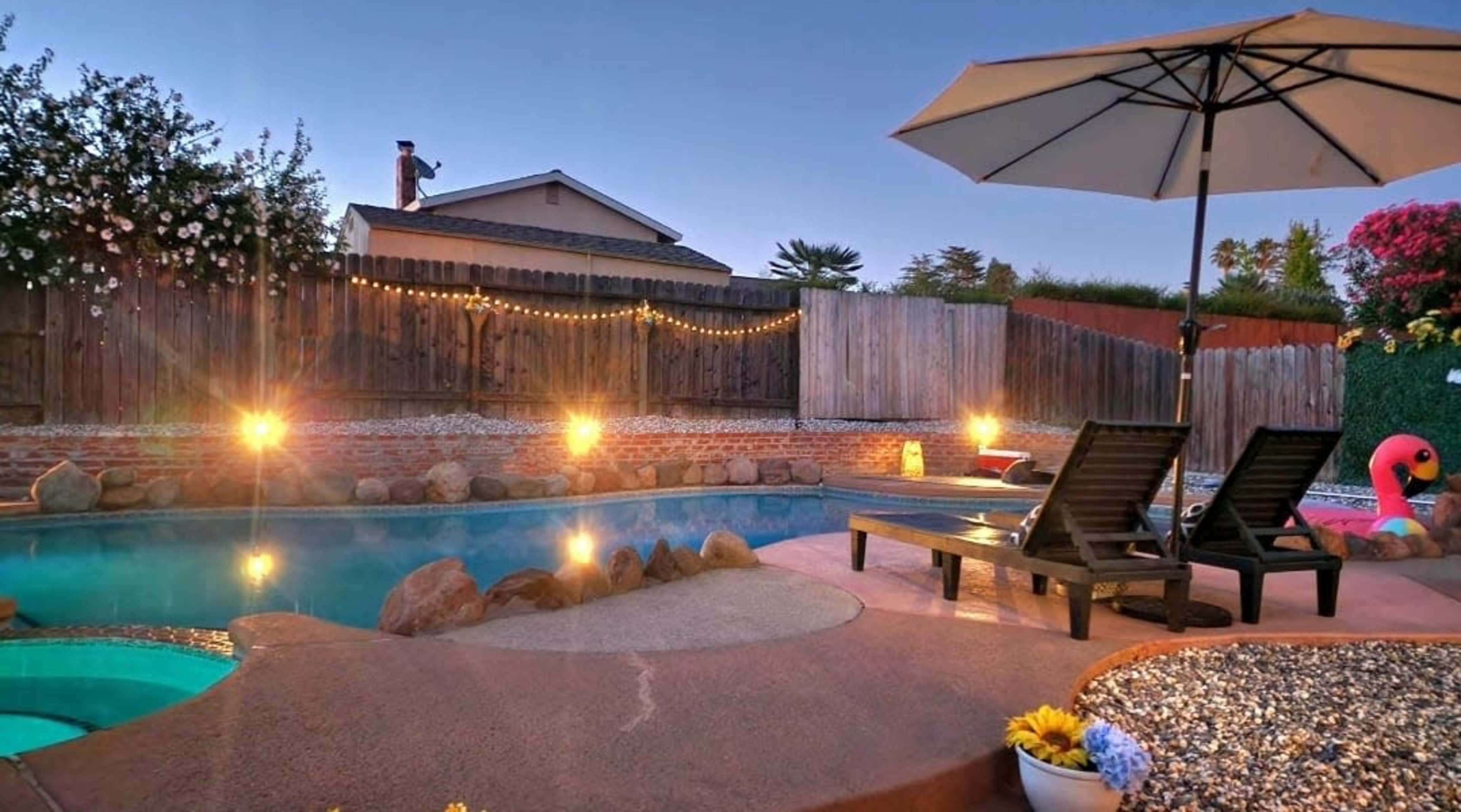 A swimming pool with lounge chairs and an umbrella is surrounded by a wooden fence, illuminated by string lights and evening lights.