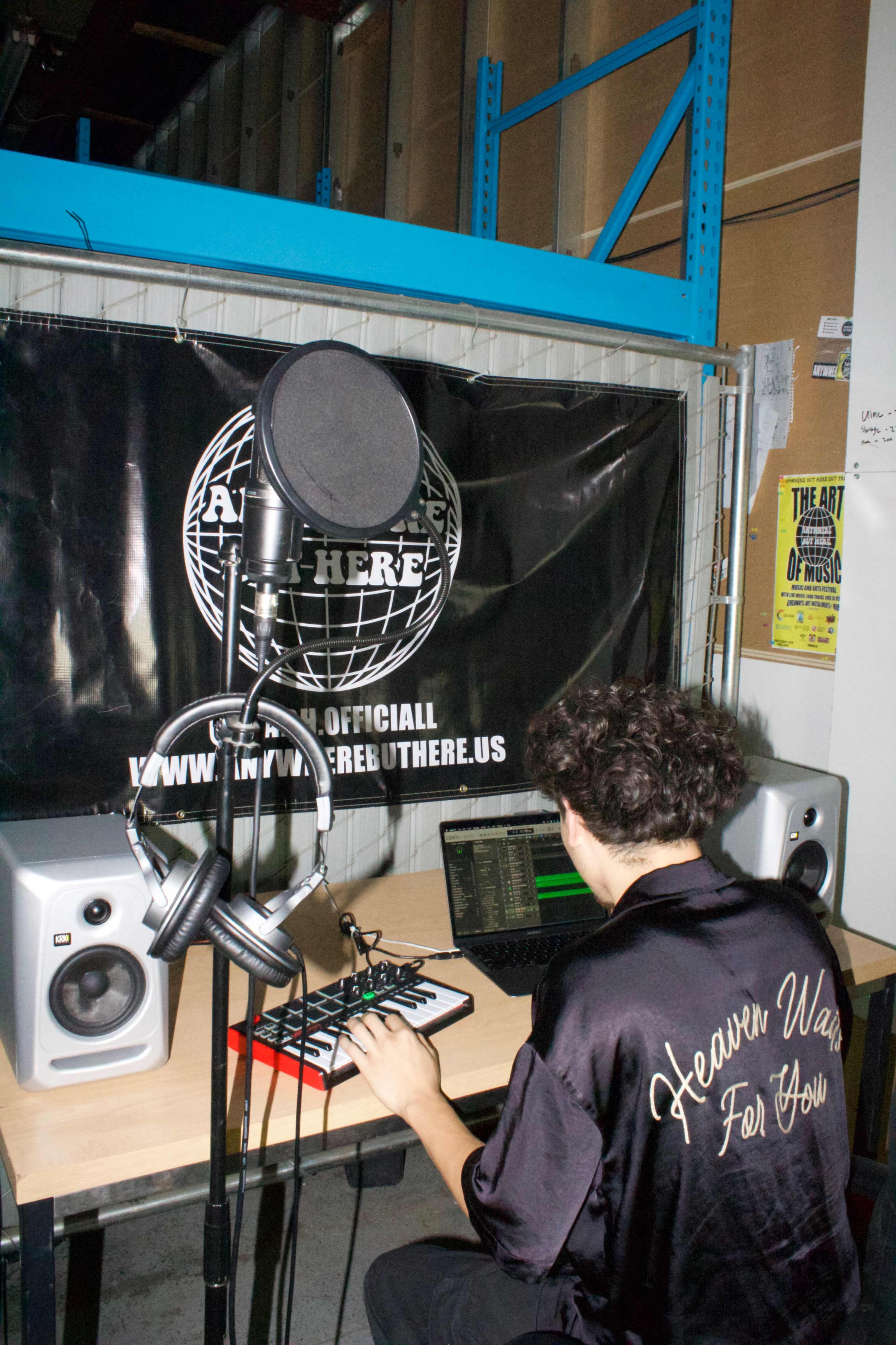 A person wearing a black satin jacket sits at a desk with music equipment, including a microphone and speakers, in front of a backdrop featuring a website banner.