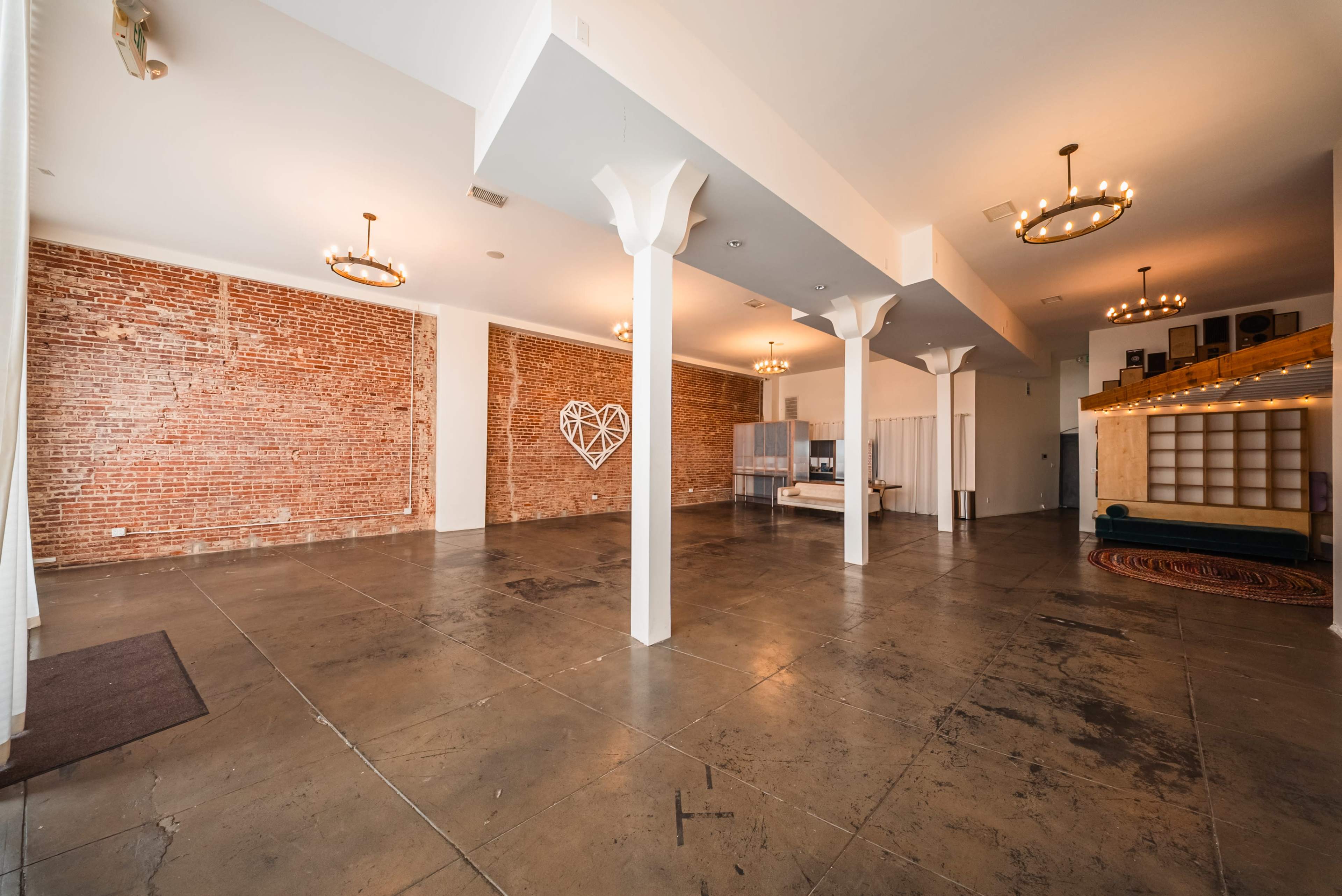The image shows a spacious, empty room with exposed brick walls, concrete flooring, and several round chandeliers hanging from the ceiling.