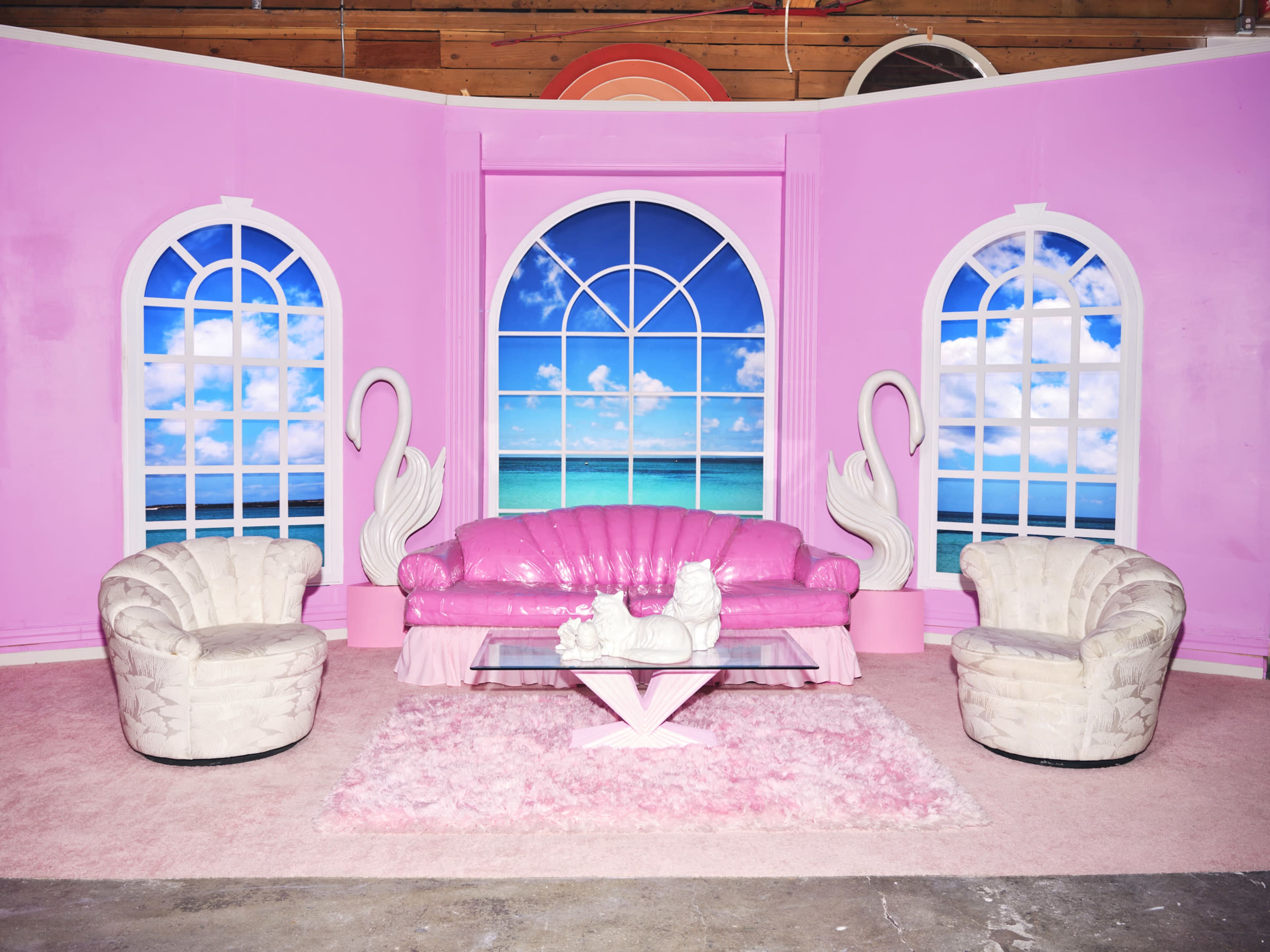 A vibrant pink room features a plush pink sofa, two white armchairs, and a glass coffee table, set against a backdrop of large windows showcasing a tropical beach scene.