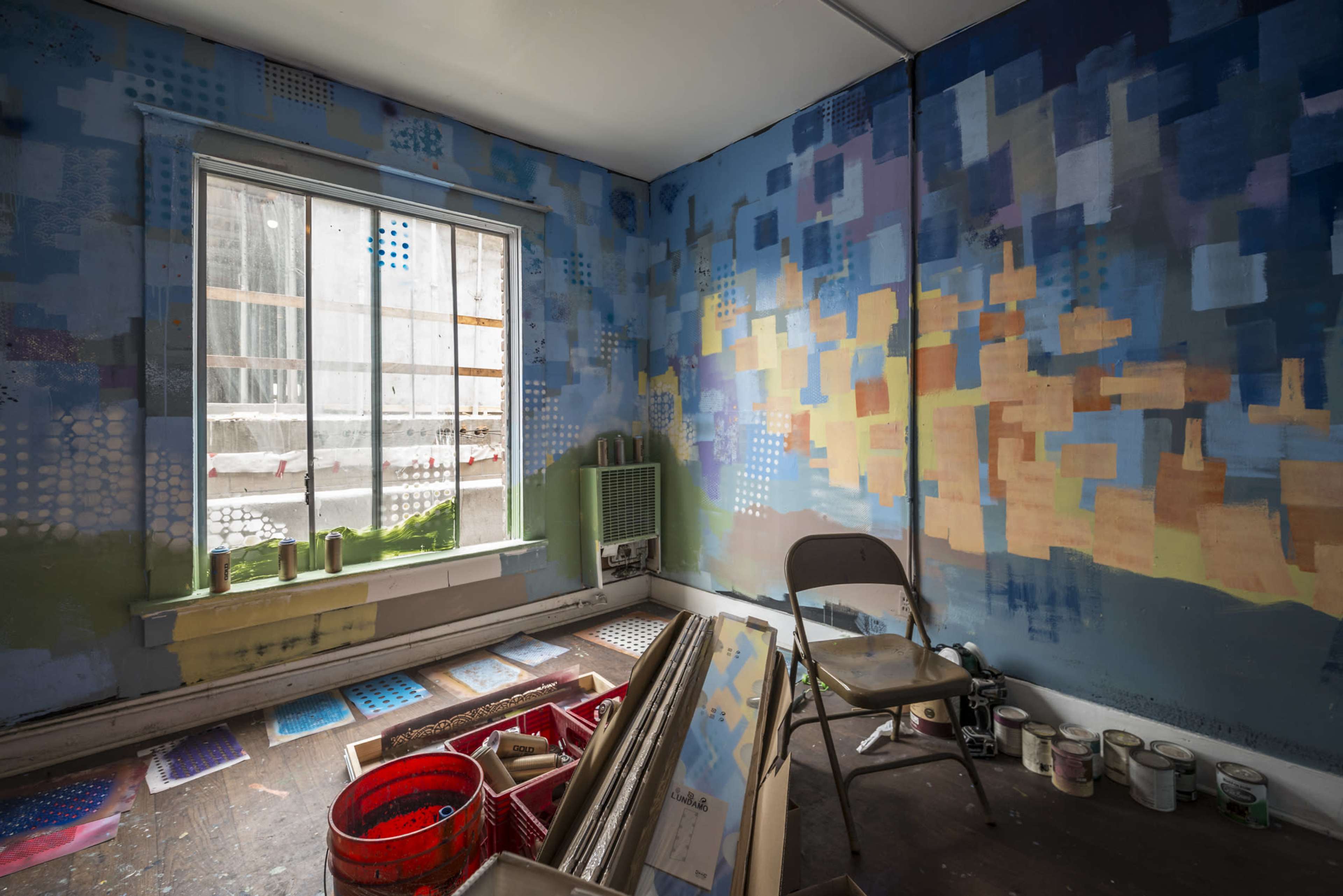 The room features colorful, abstract wall art with a window revealing an outdoor view and a chair placed beside various art supplies and containers.