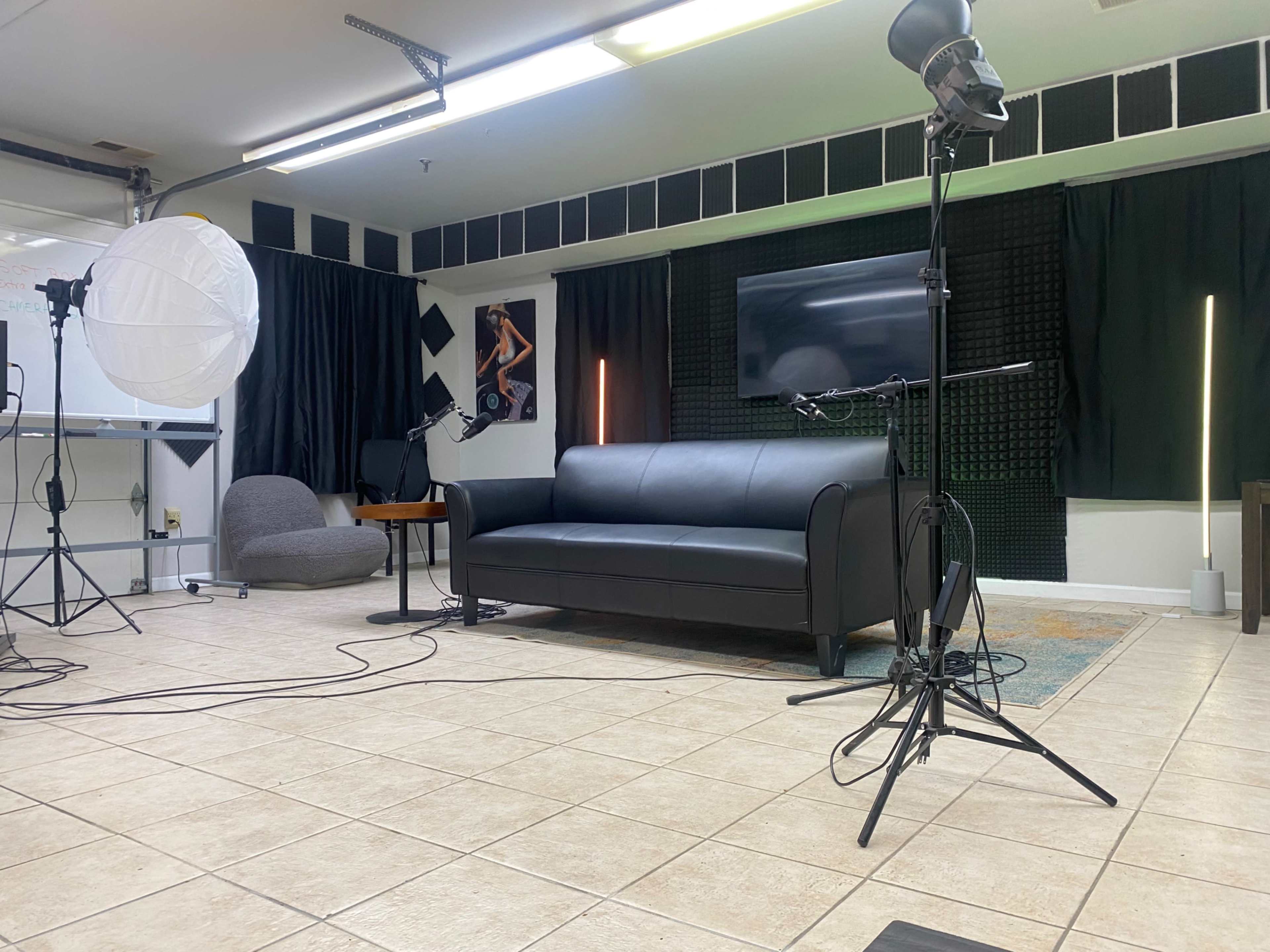 A studio setup with a black sofa, a standing microphone on a tripod, and lighting equipment against a backdrop of black curtains and a television.