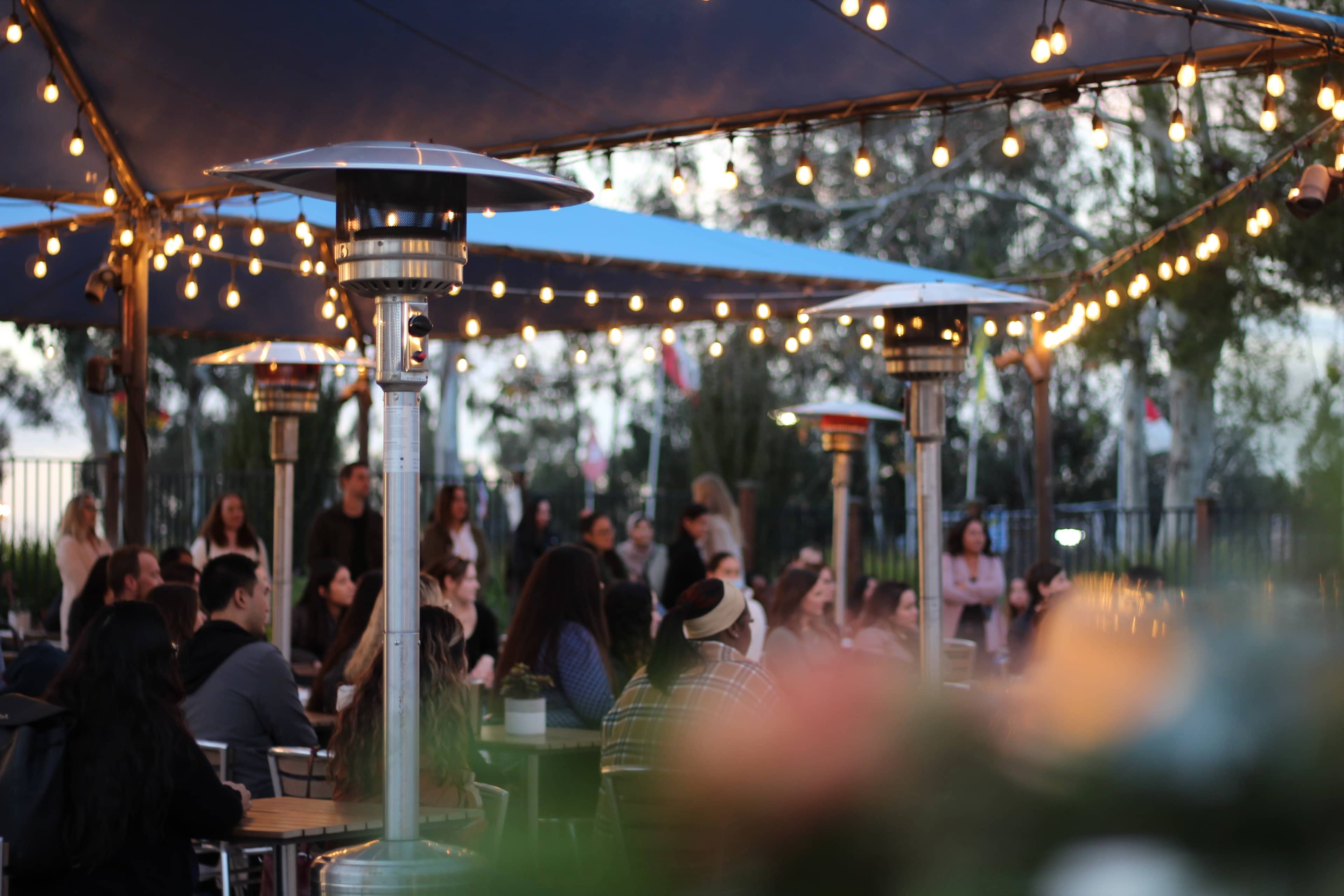 A lively outdoor gathering is illuminated by string lights and heat lamps, with people seated at tables beneath a large tent.