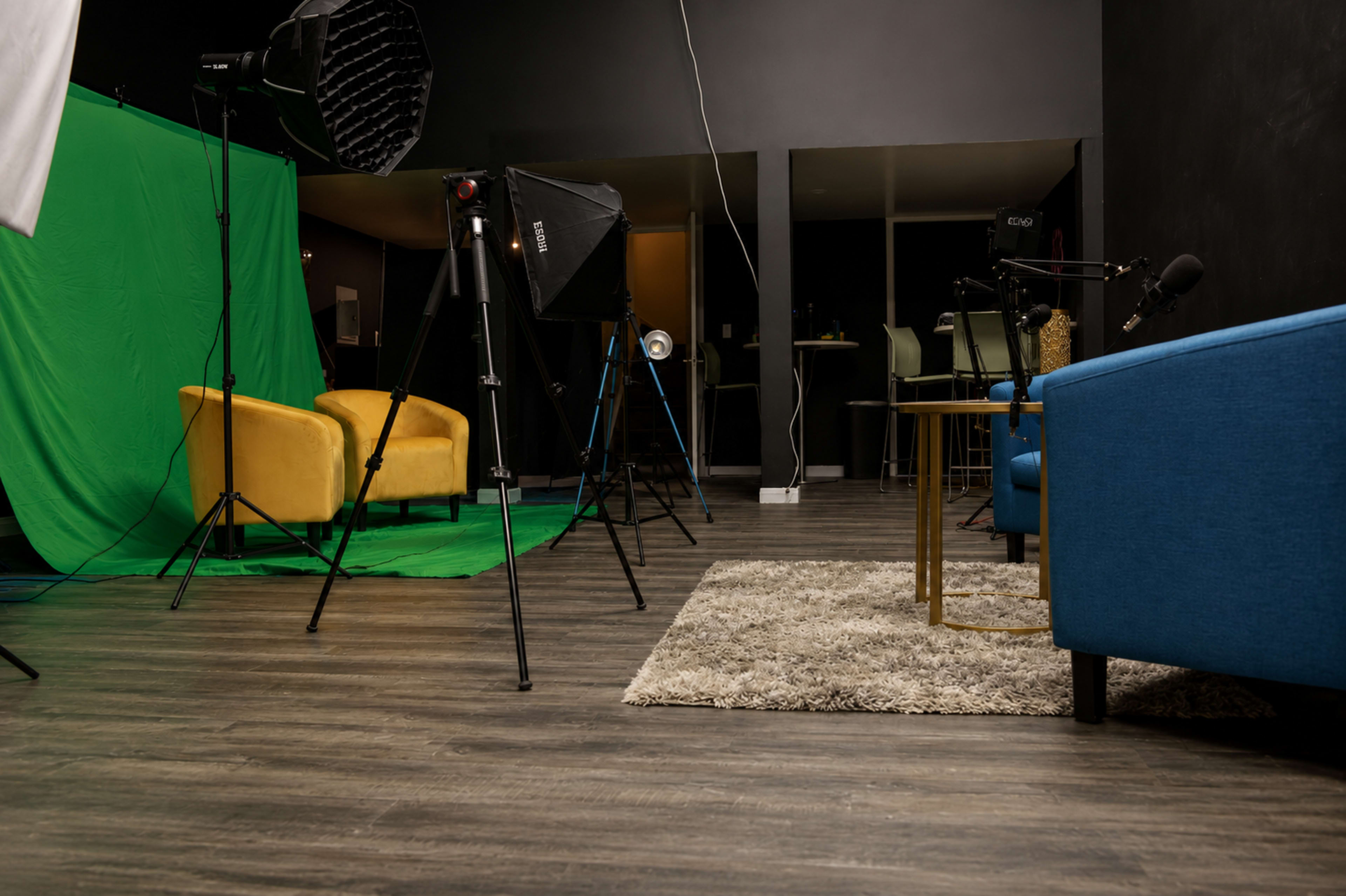 The image shows a studio setup featuring a green screen, various lighting equipment, and two yellow chairs positioned in front of a blue couch on a rug.
