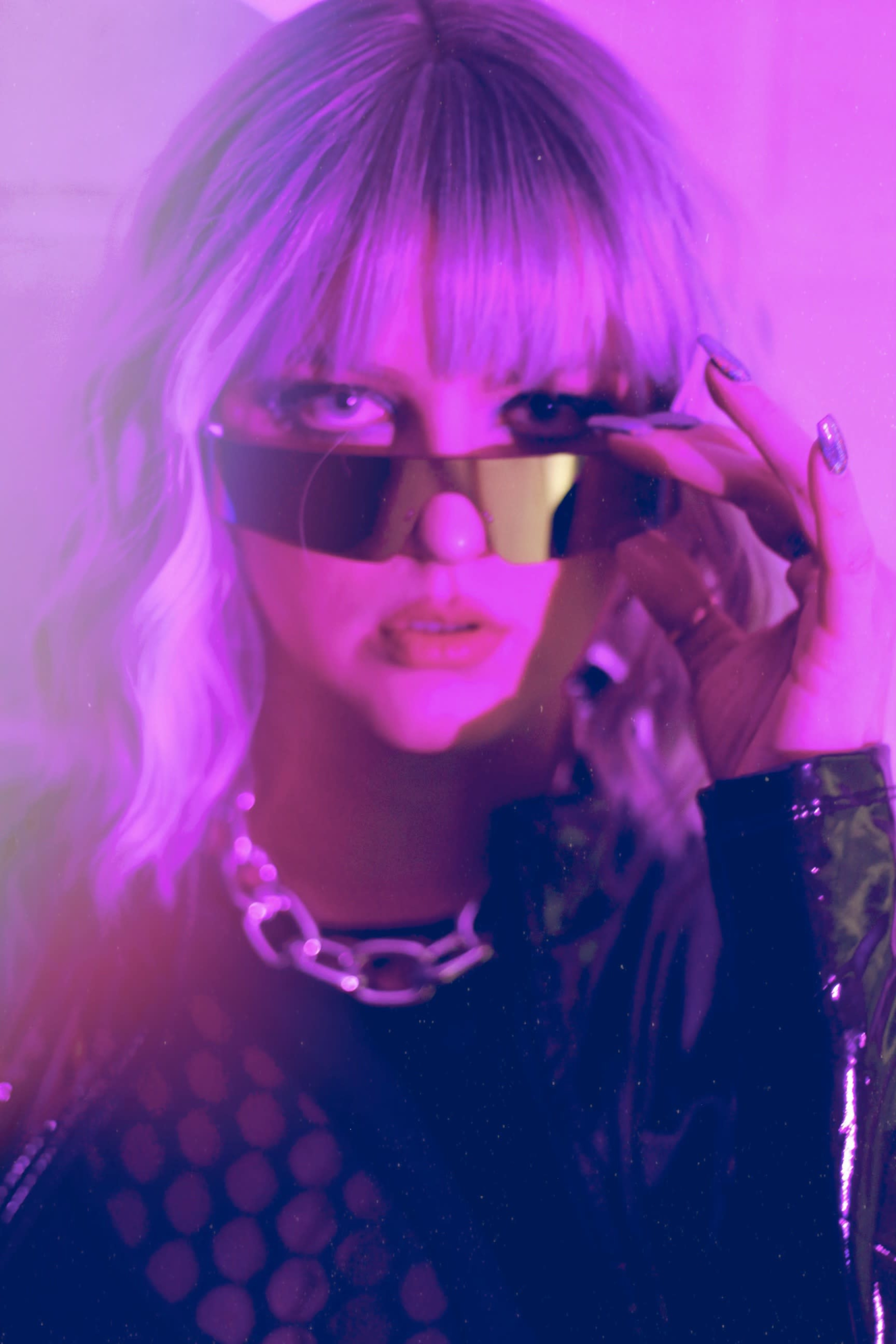 A person with light purple hair wearing sunglasses and a chain necklace gazes at the camera against a colorful background.