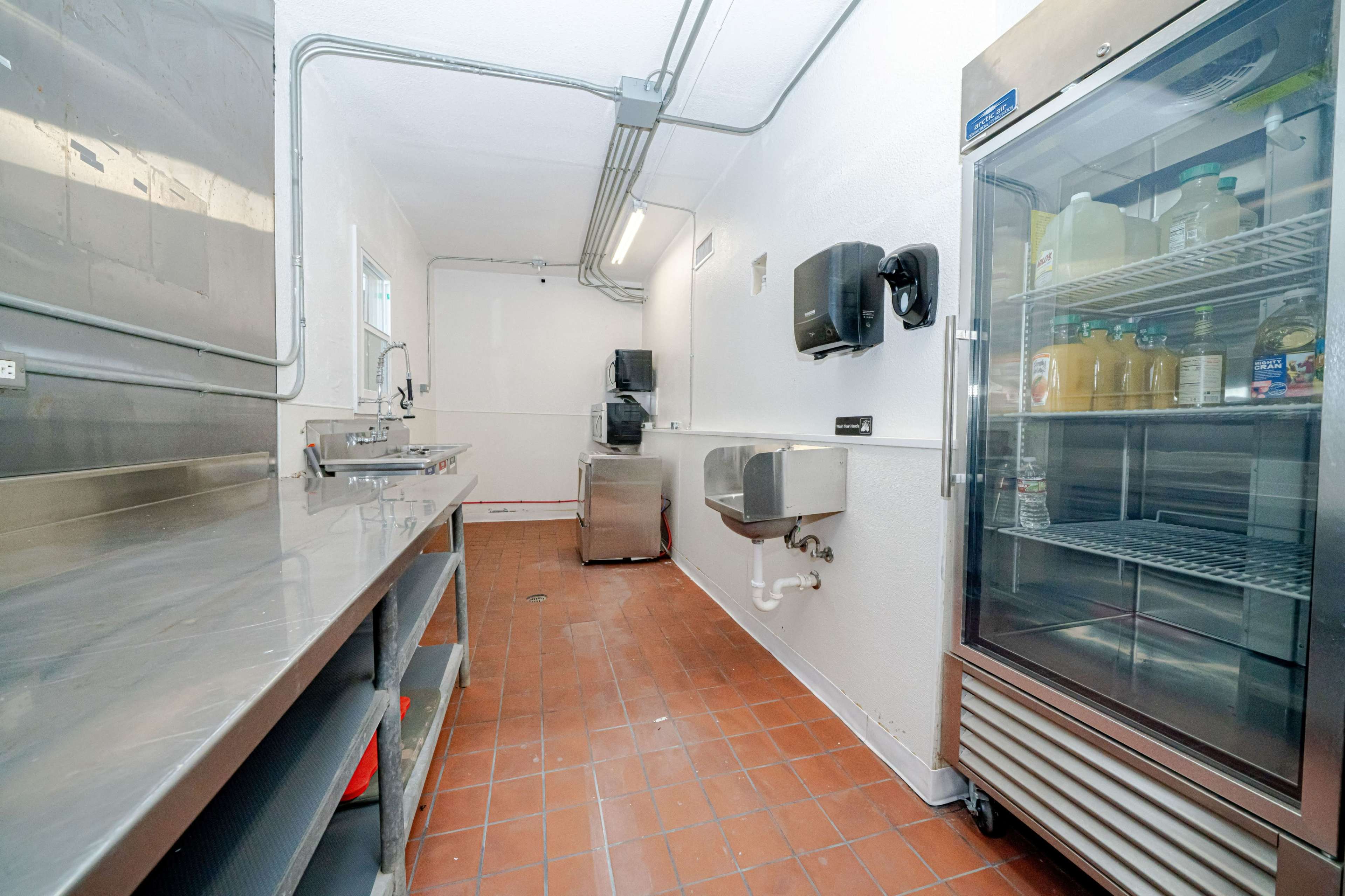 The image shows a clean commercial kitchen space with stainless steel countertops, a sink, and a refrigerator along the wall.