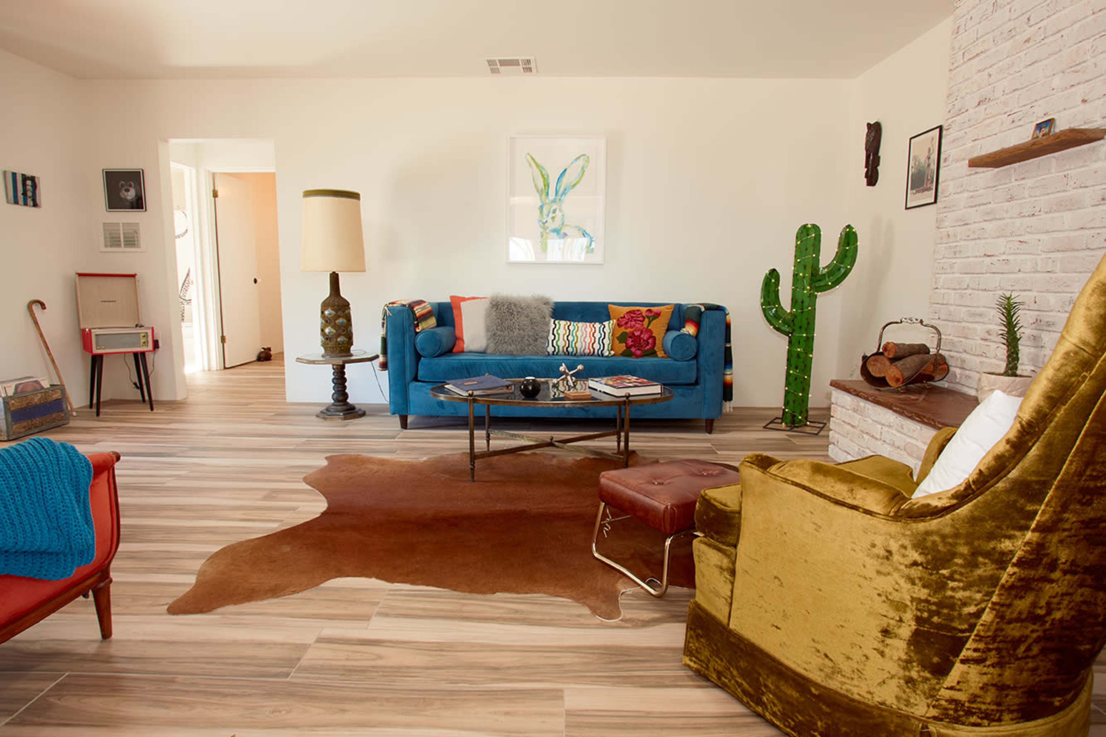 The image shows a modern living room featuring a blue sofa, an armchair with a gold fabric, a coffee table on a cowhide rug, and a decorative cactus in a corner.