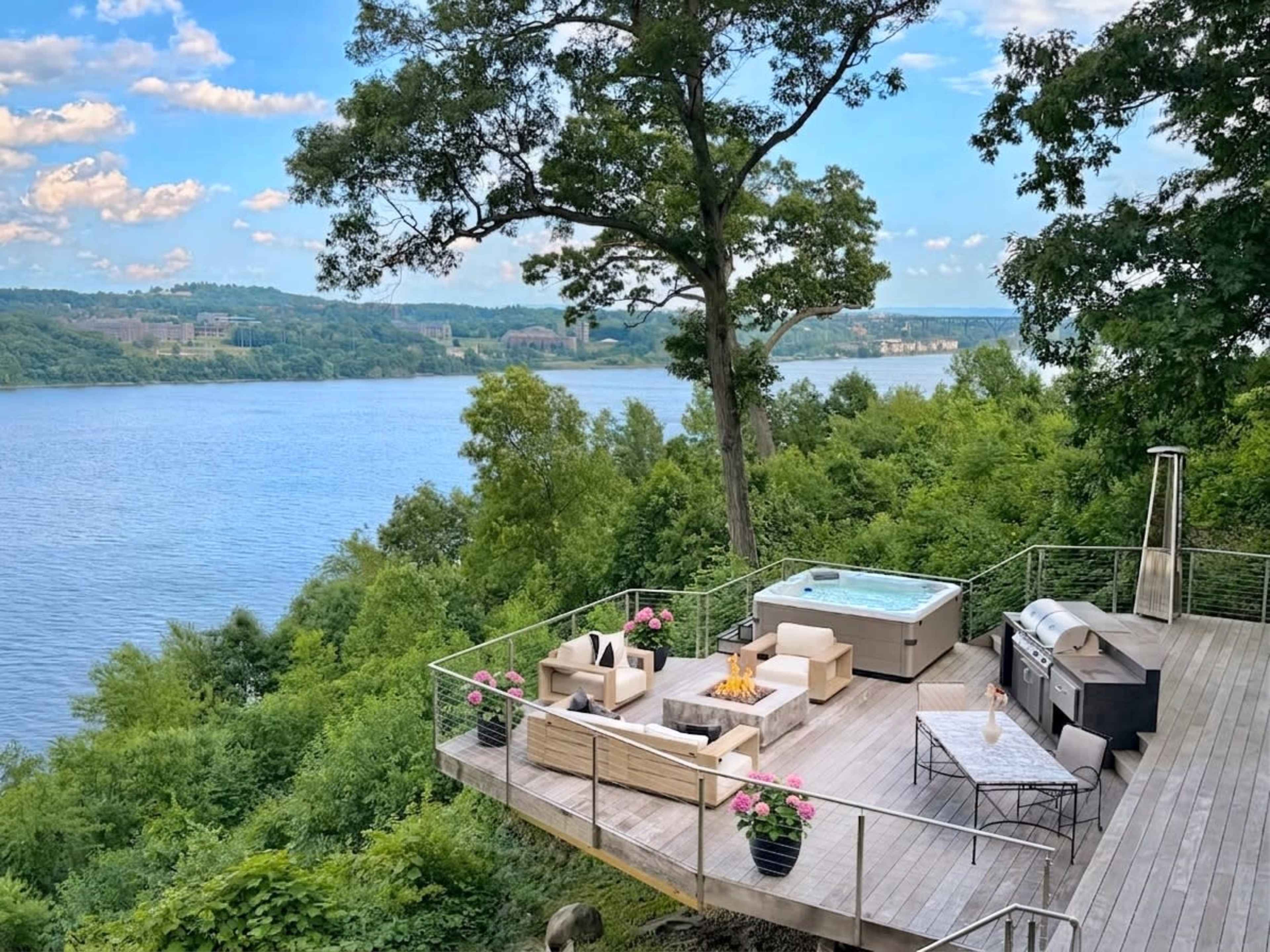A wooden deck overlooking a river features comfortable outdoor seating, a hot tub, a barbecue grill, and potted plants.
