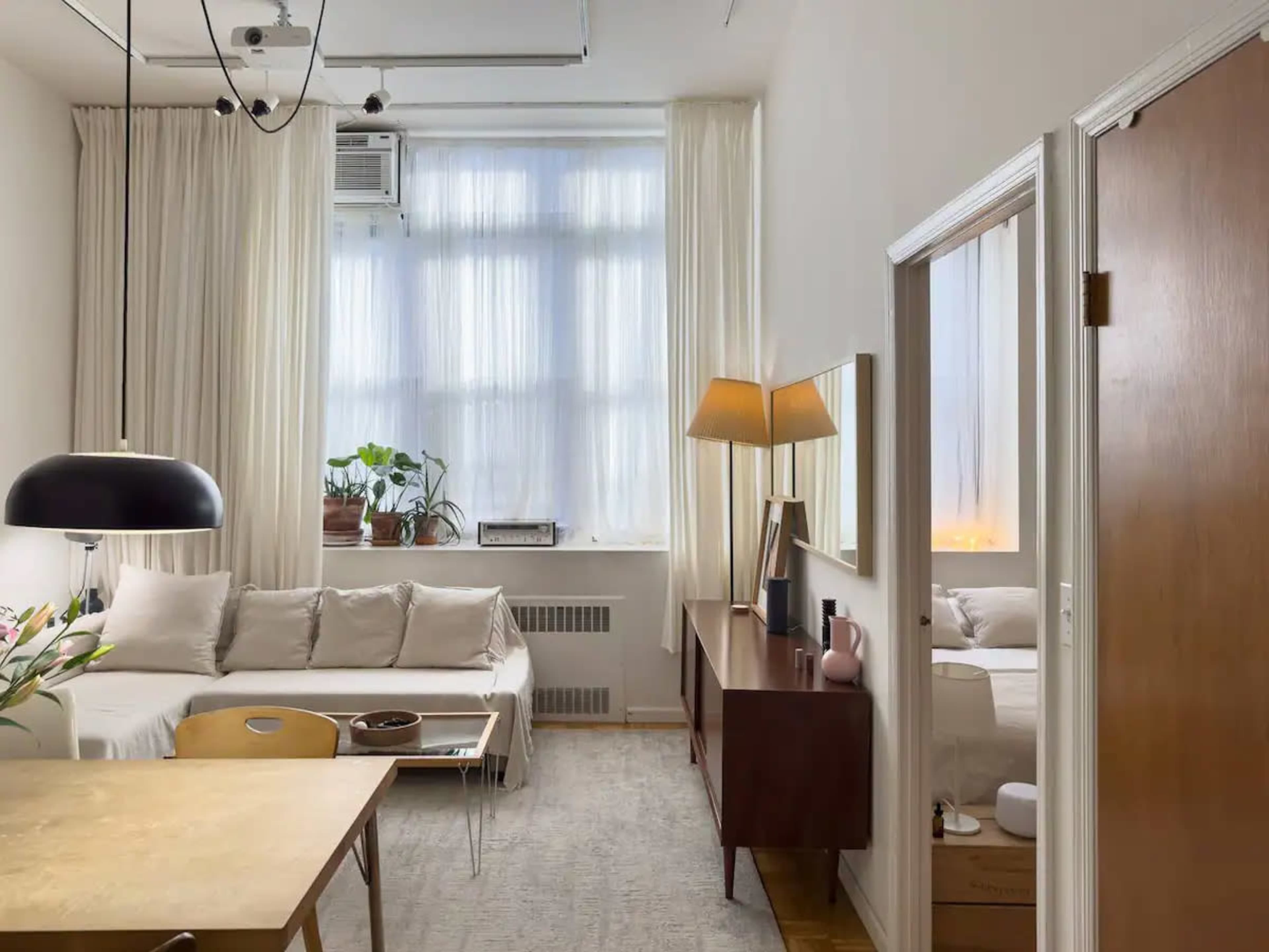 The image shows a bright, modern living space featuring a sofa, a dining table, and a mid-century style sideboard, with large windows covered by sheer curtains.