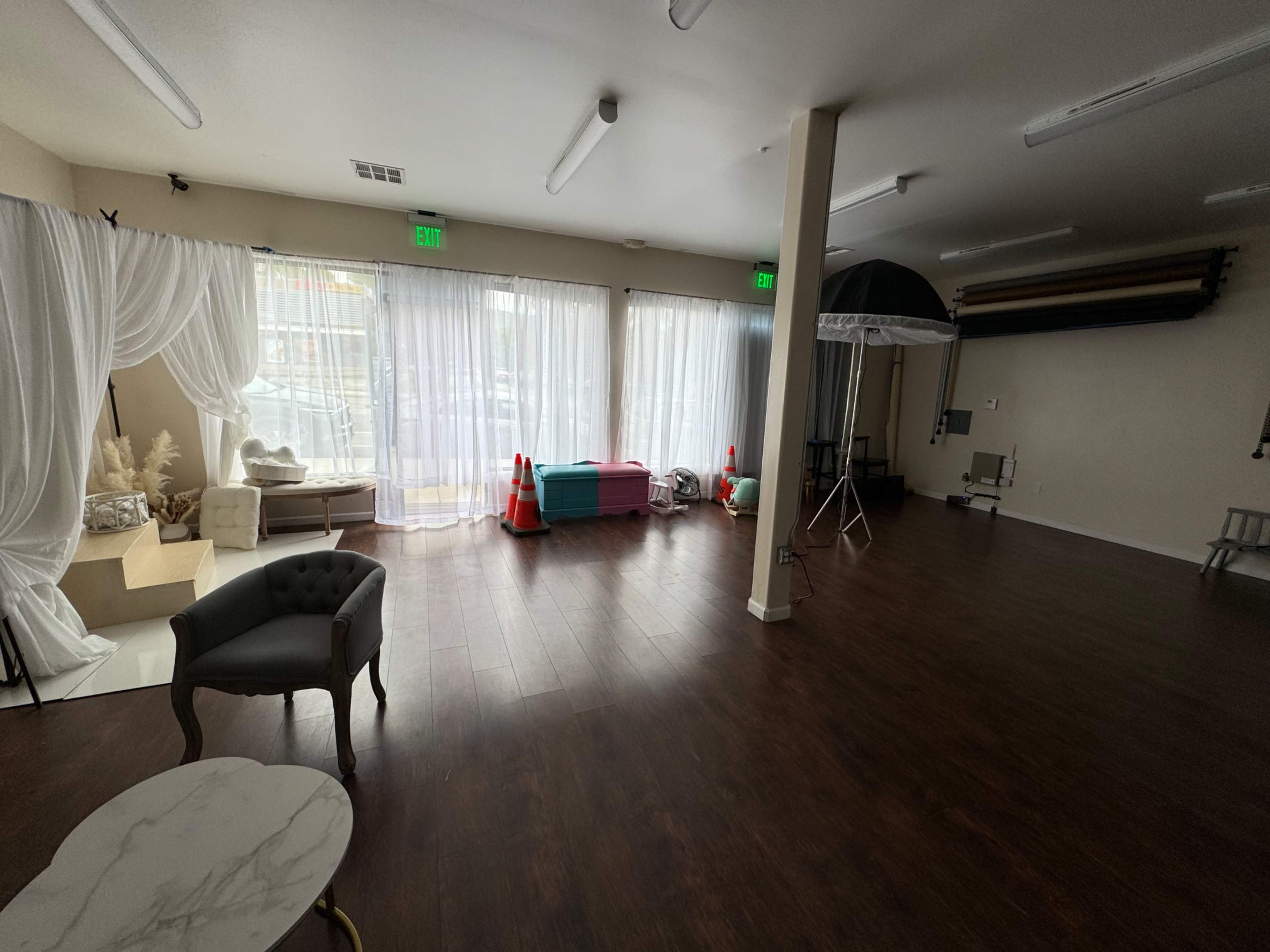An interior space with wooden flooring, large windows covered by sheer curtains, and a mix of furniture including a chair and decorative elements.