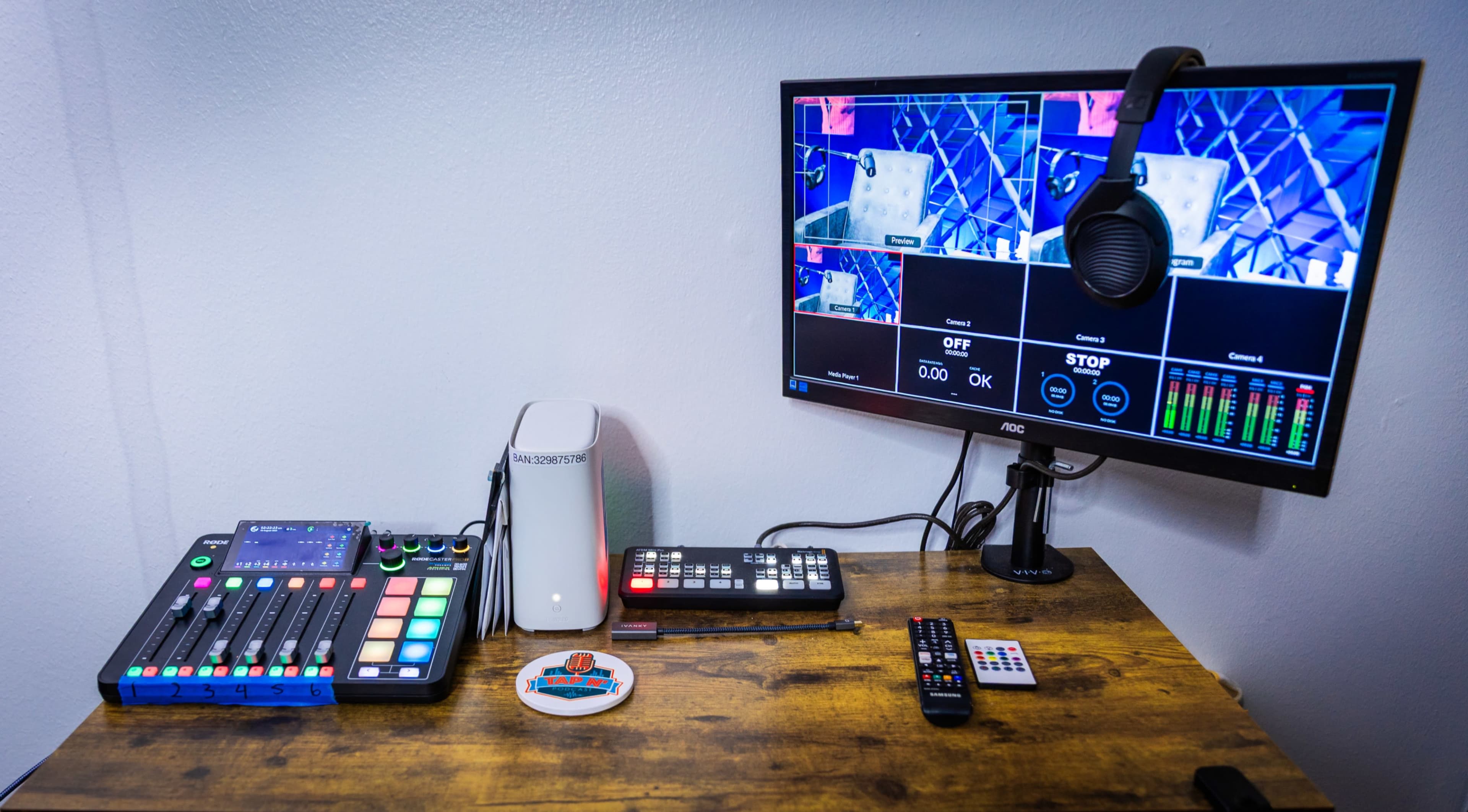 The image shows a desk with a digital audio mixer, a computer monitor displaying video feeds, and a pair of headphones, all arranged neatly on a wooden surface.