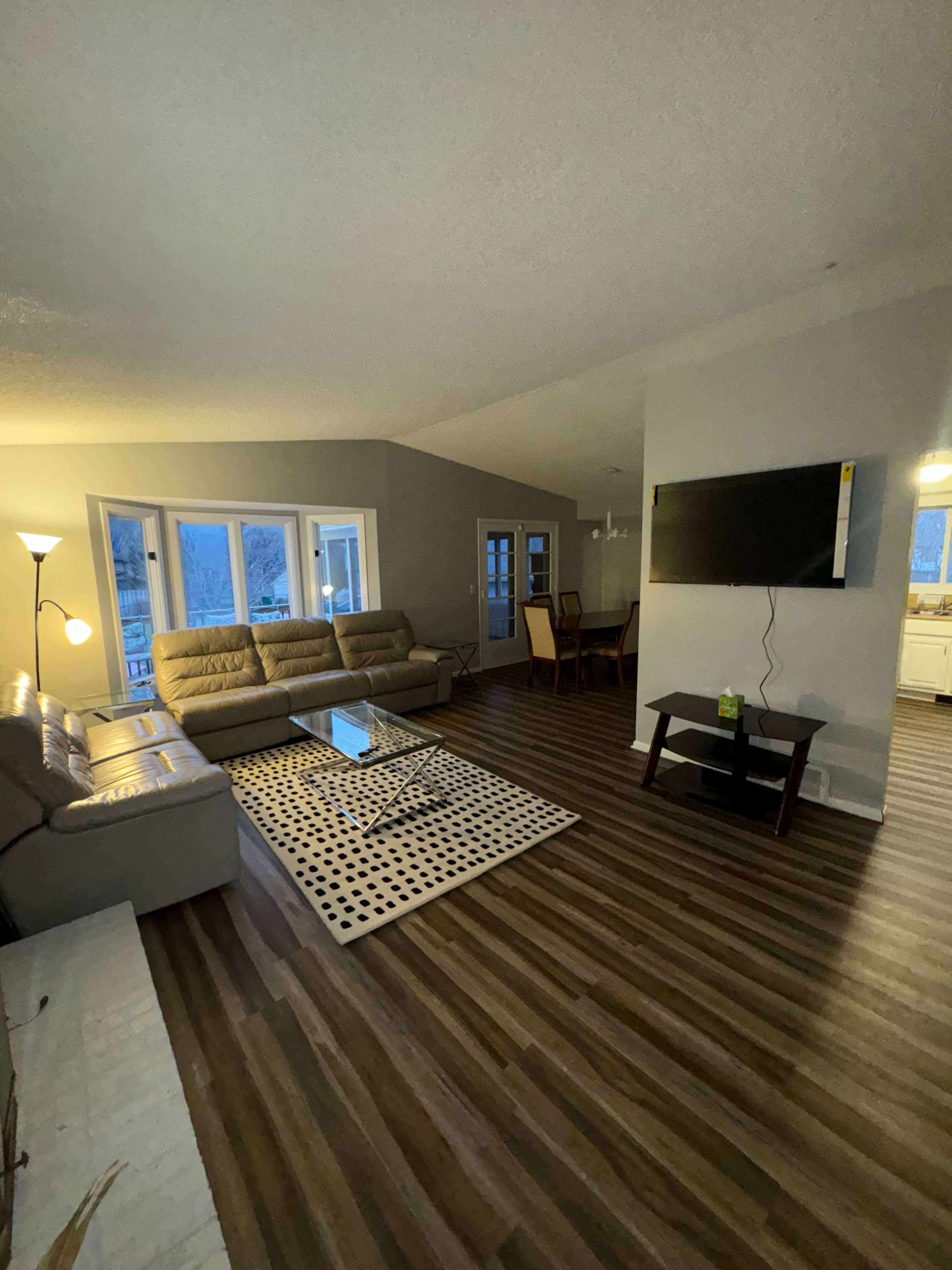 A modern living room with a sectional sofa, a coffee table on a patterned rug, a wall-mounted television, and dining area visible in the background.