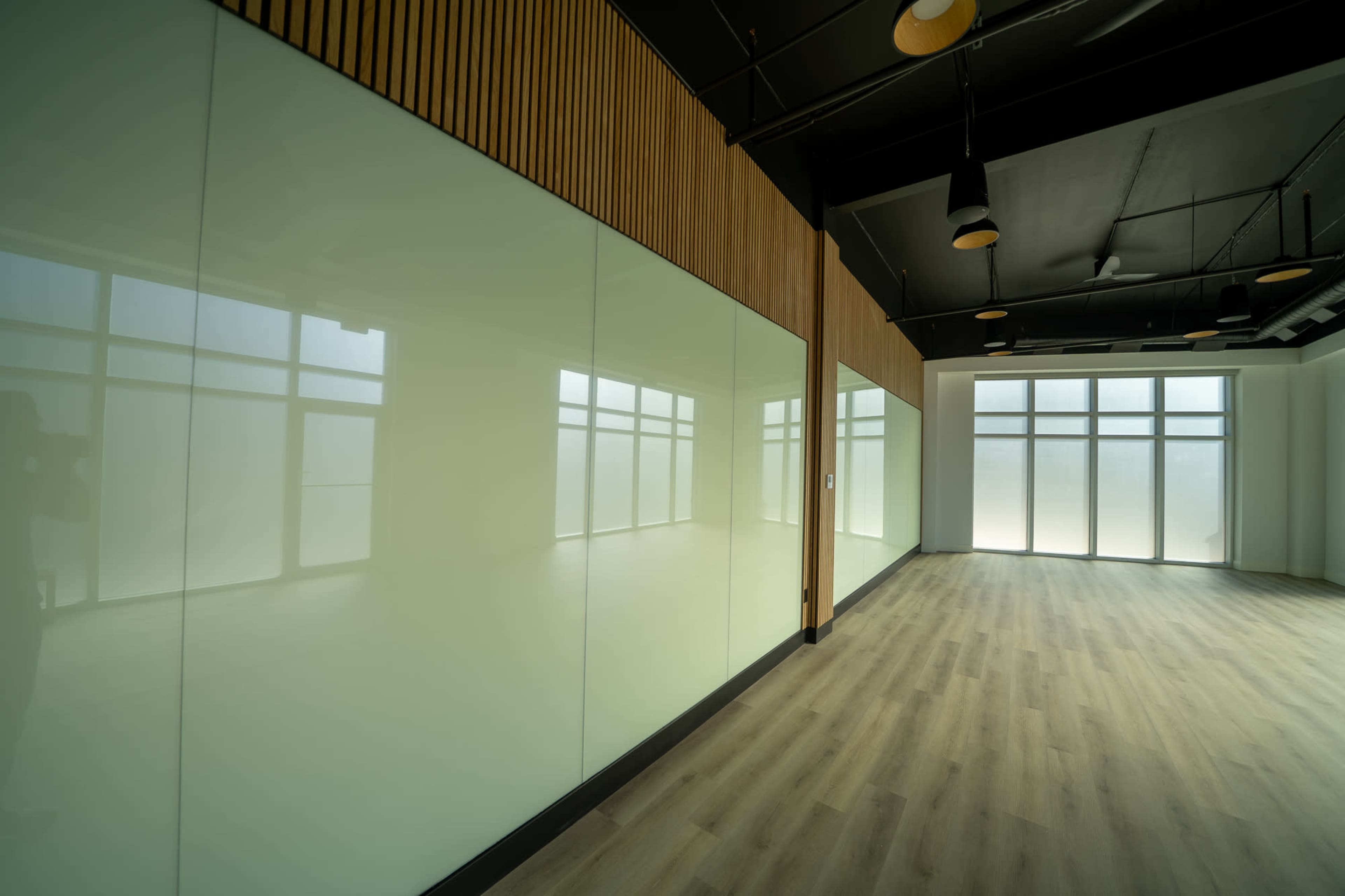 The image shows an empty, modern office space with large glass walls and wooden accents, featuring light-colored flooring.