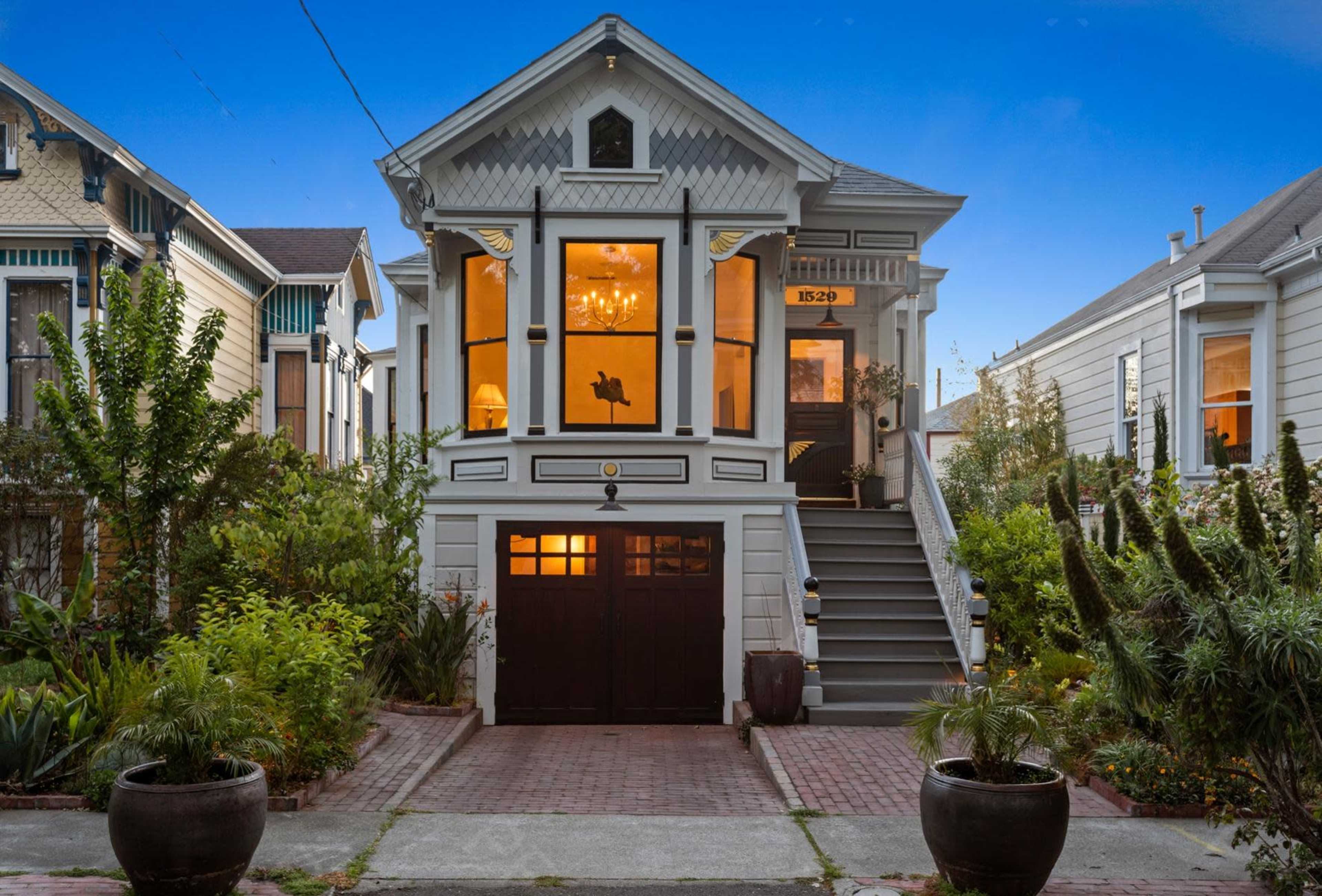 A Victorian-style house with a dark garage door and large windows is surrounded by greenery and features a set of stairs leading to the front entrance.