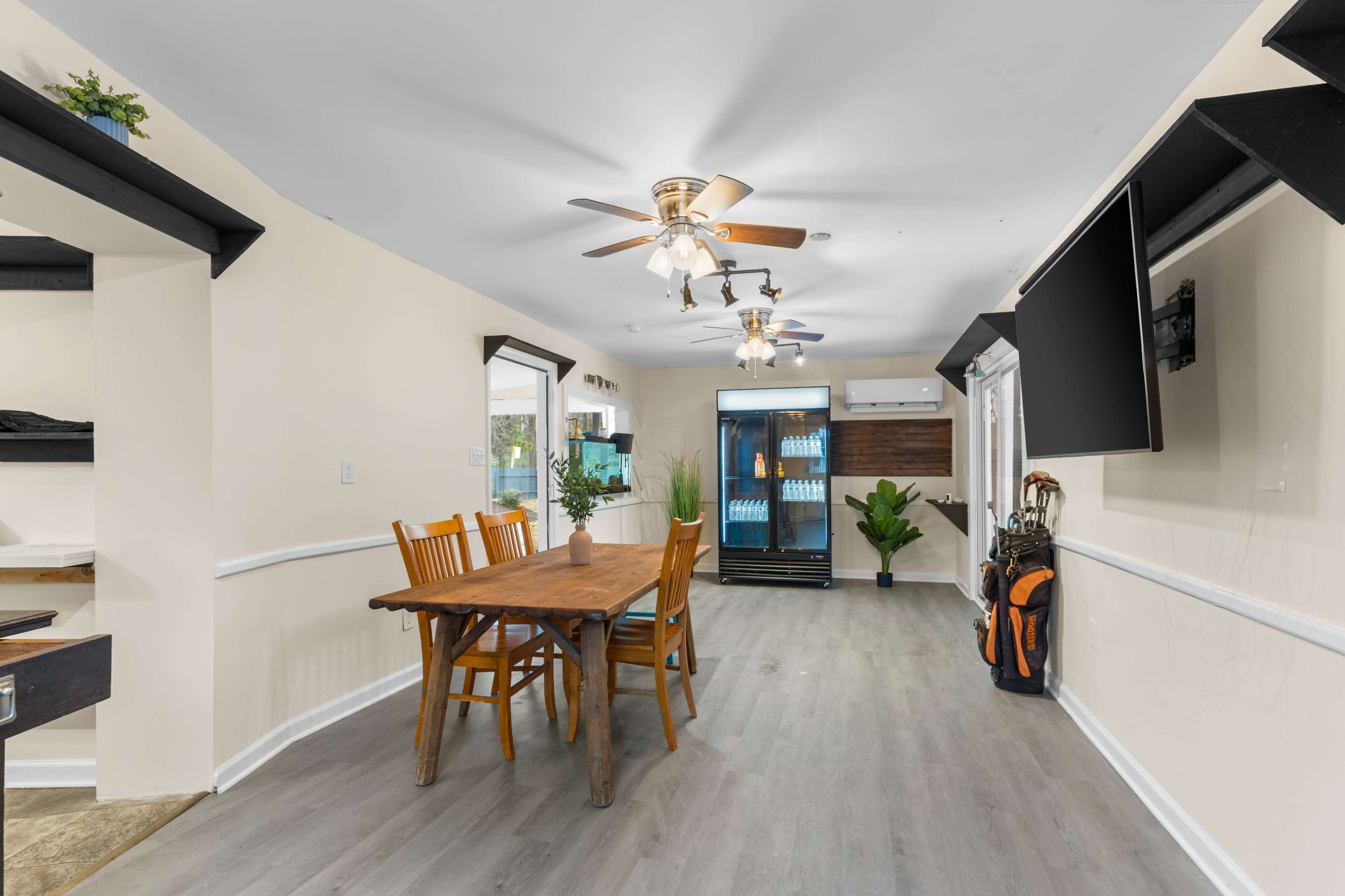 The image shows a dining area with a wooden table and chairs, surrounded by light-colored walls, a television mounted on one side, and a display refrigerator in the background.