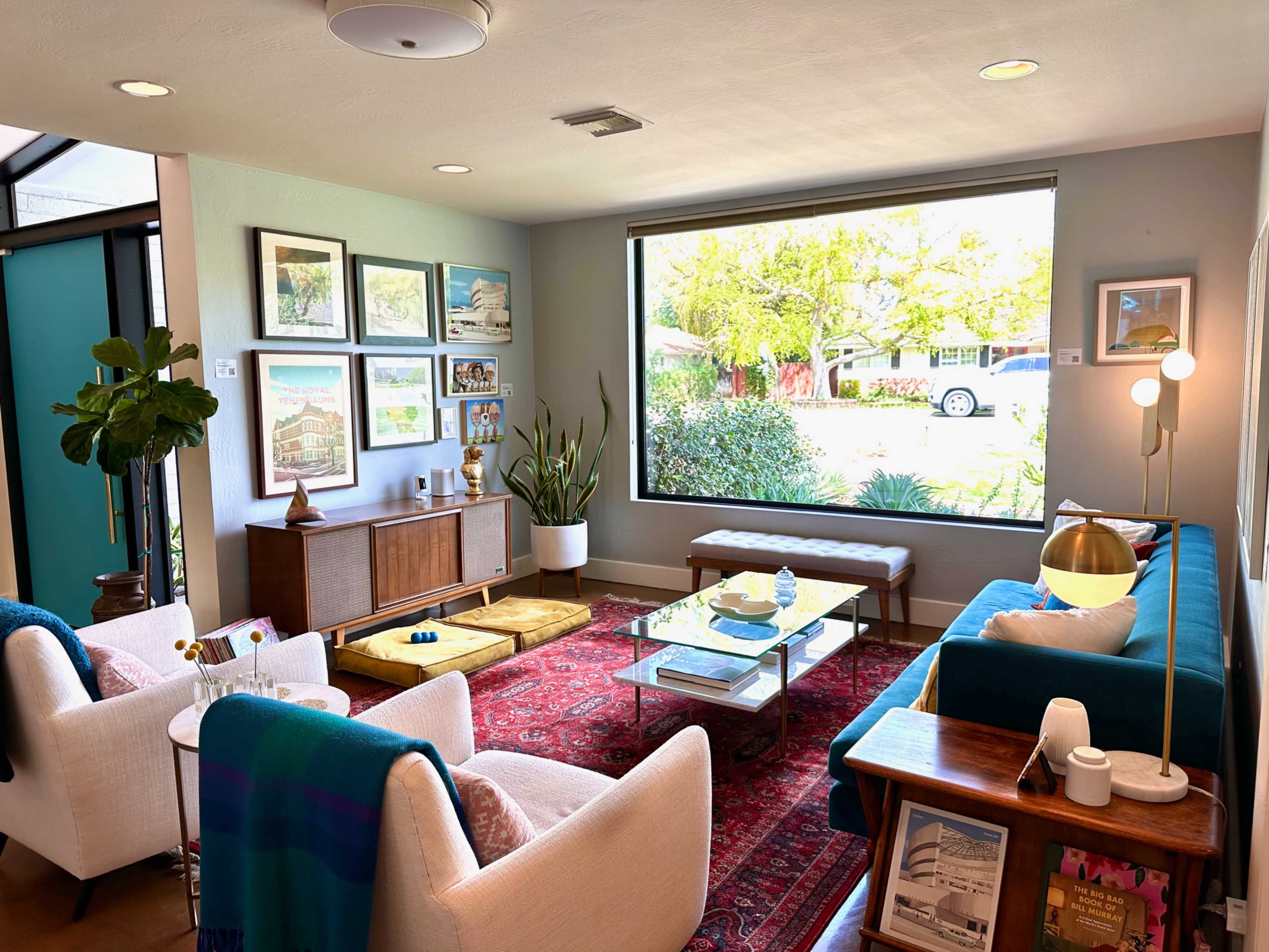A bright living room features a large window overlooking a garden, with comfortable seating, art on the walls, and a coffee table in the center.
