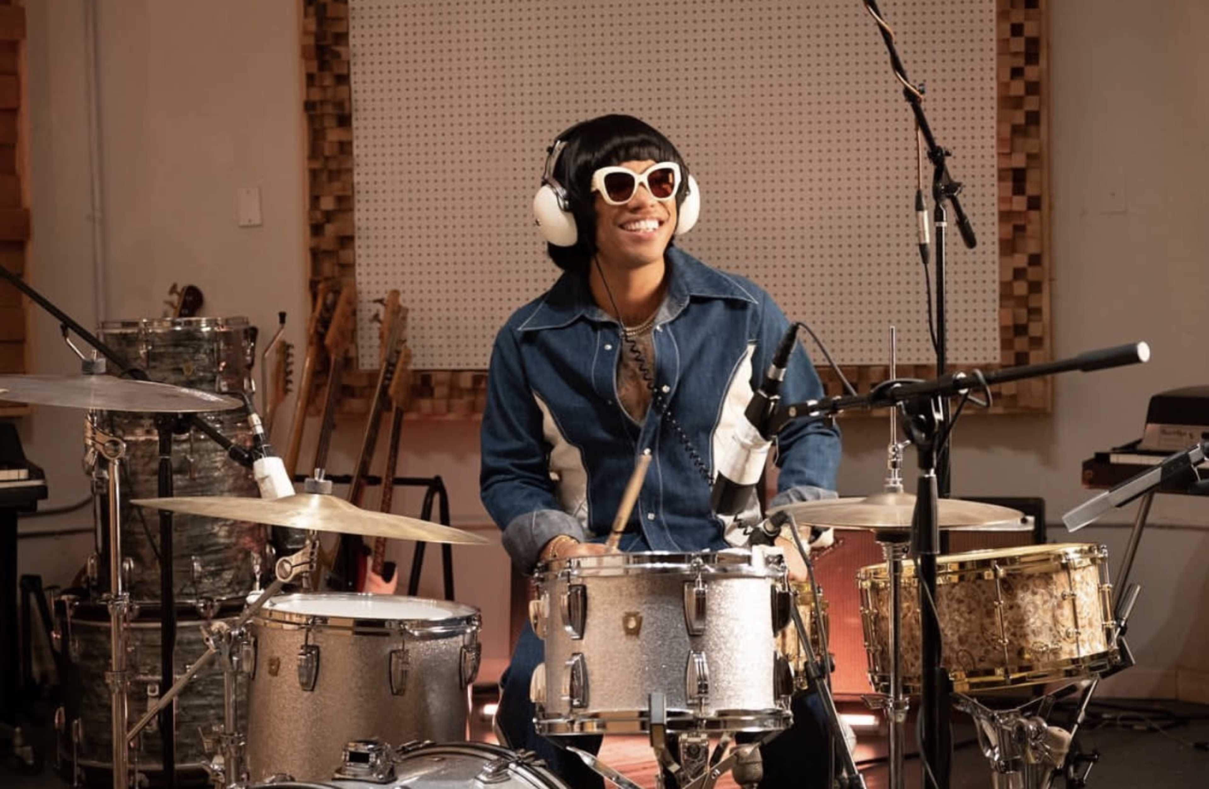 A person wearing sunglasses and headphones plays the drums in a music studio surrounded by instruments.